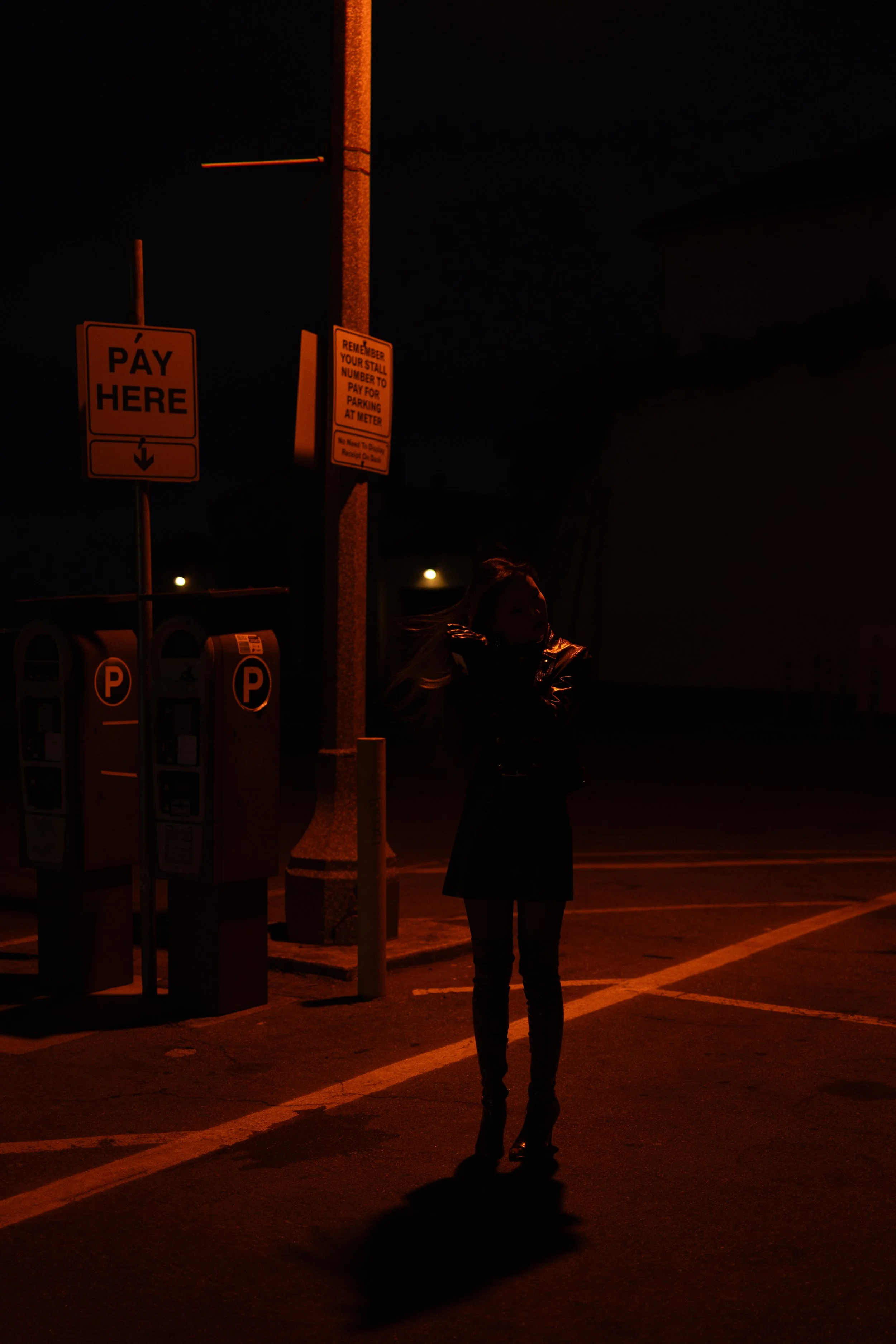 A woman standing alone in a parking lot at night, illuminated by nearby streetlights, with parking meters and signs nearby.