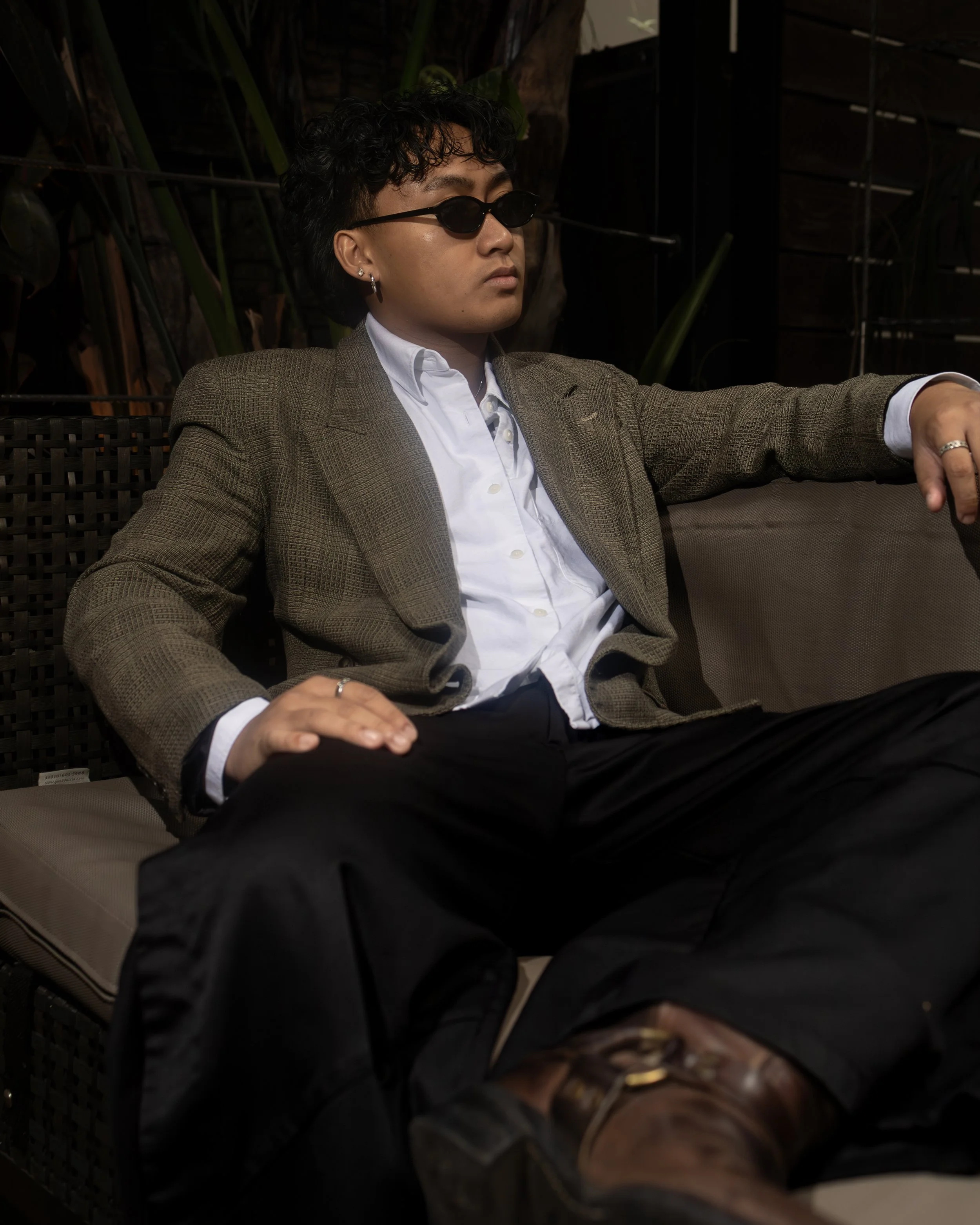 A man wearing a brown plaid blazer and white shirt, with sunglasses and silver hoop earrings, sitting on a dark woven outdoor chair with a view of plants and a wooden fence in the background.
