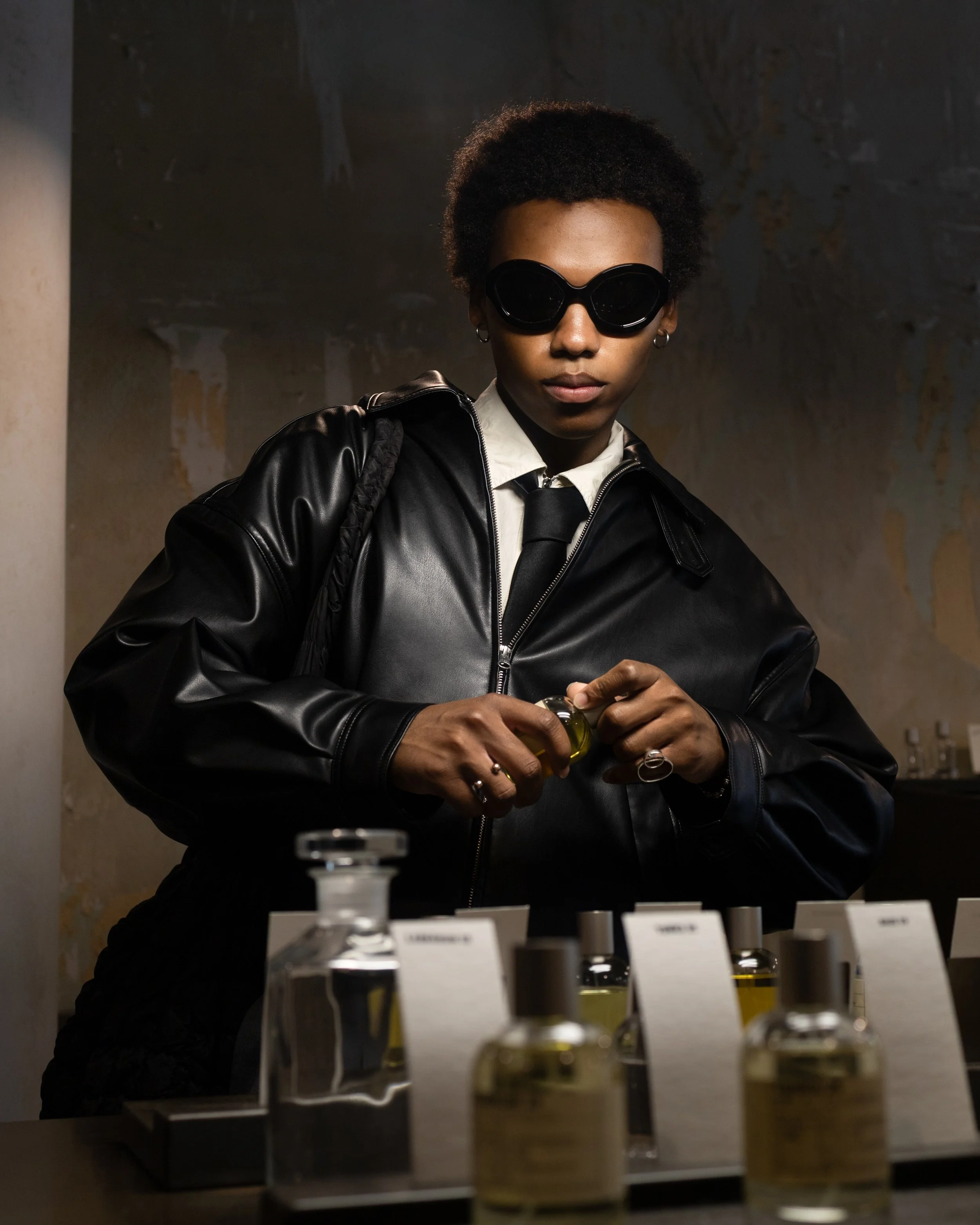 A woman with short curly hair wearing sunglasses, a white shirt, black tie, and black leather jacket, standing behind a display of small bottles of essential oils or similar products in a dimly lit environment.