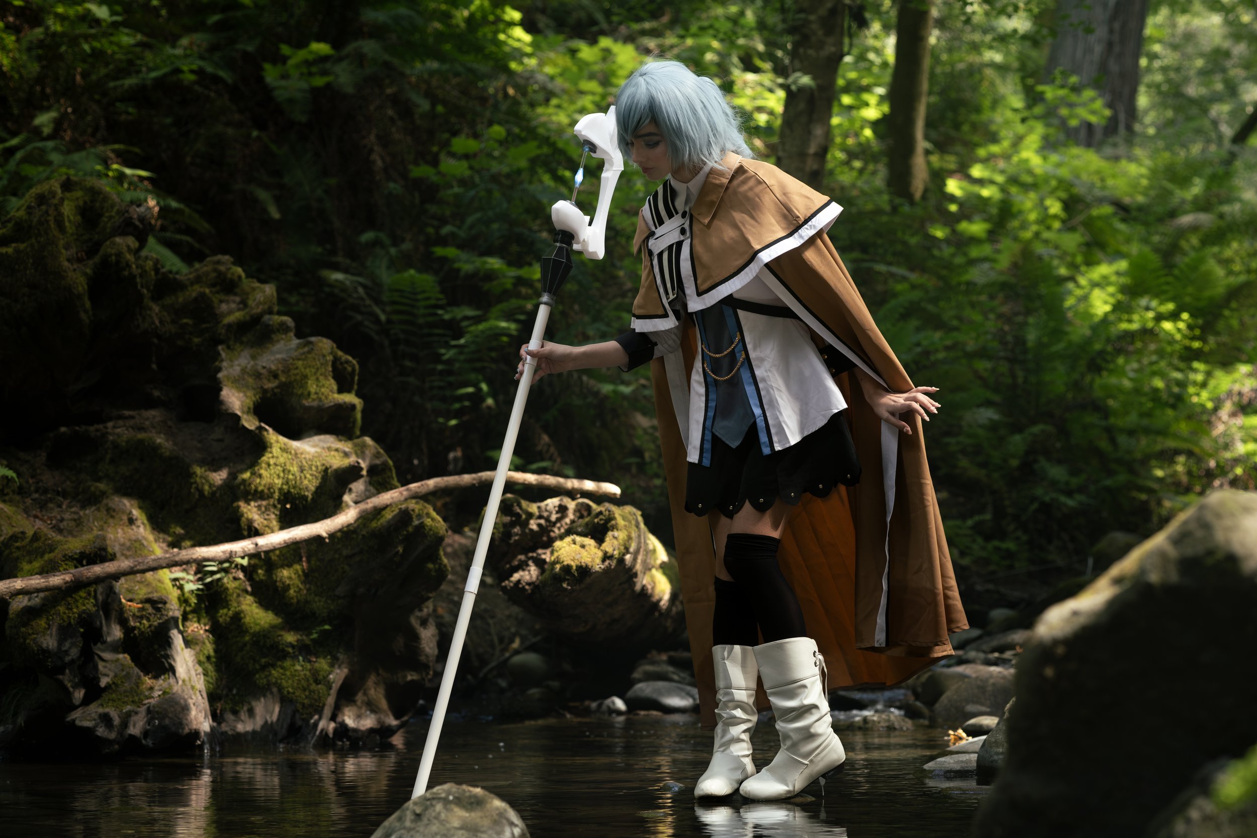 Person dressed in fantasy costume with white wig, brown and white cape, and white boots, standing in a forest stream and holding a staff.