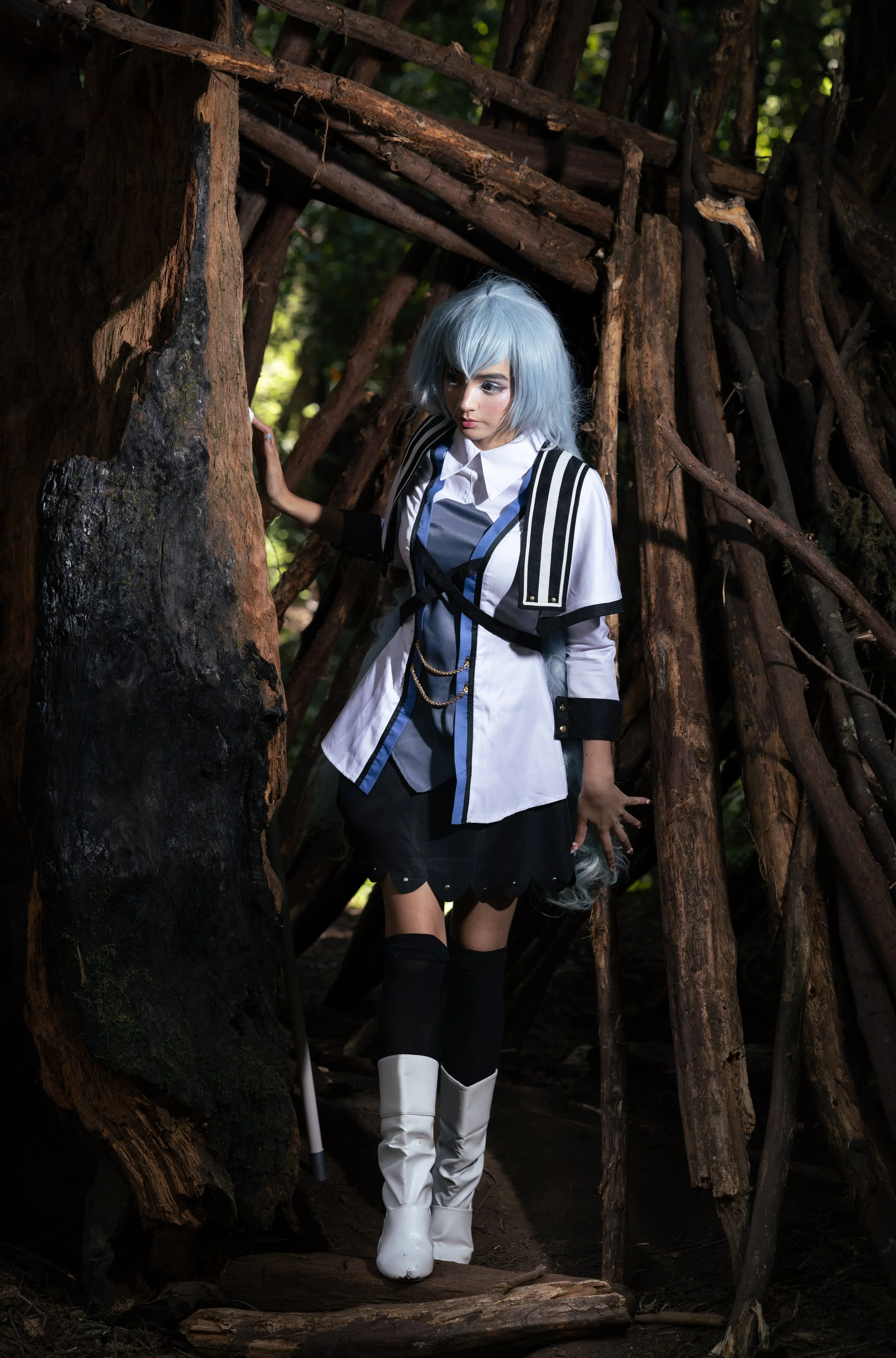 A woman with silver hair and a fantasy-inspired outfit stands in a forest, surrounded by large, twisted wooden branches and trees.