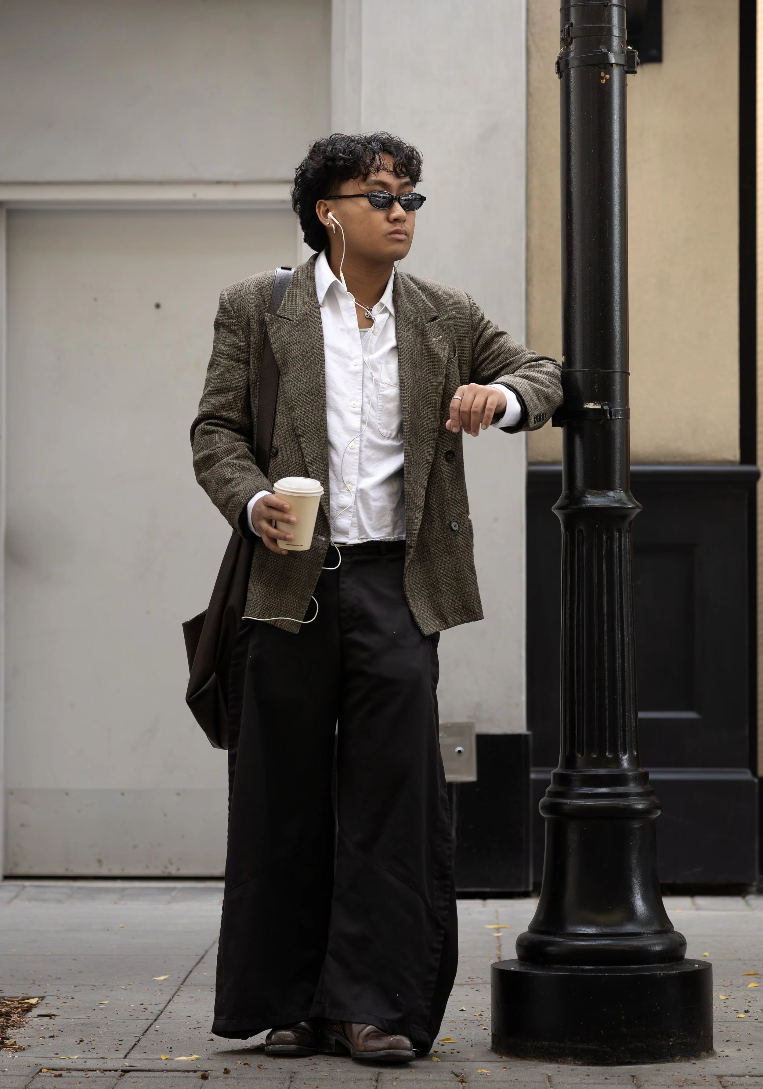 A person with curly hair wearing sunglasses, earbuds, a brown plaid blazer, white shirt, and black pants standing on a city sidewalk, looking at a black lamppost, holding a takeout coffee cup.