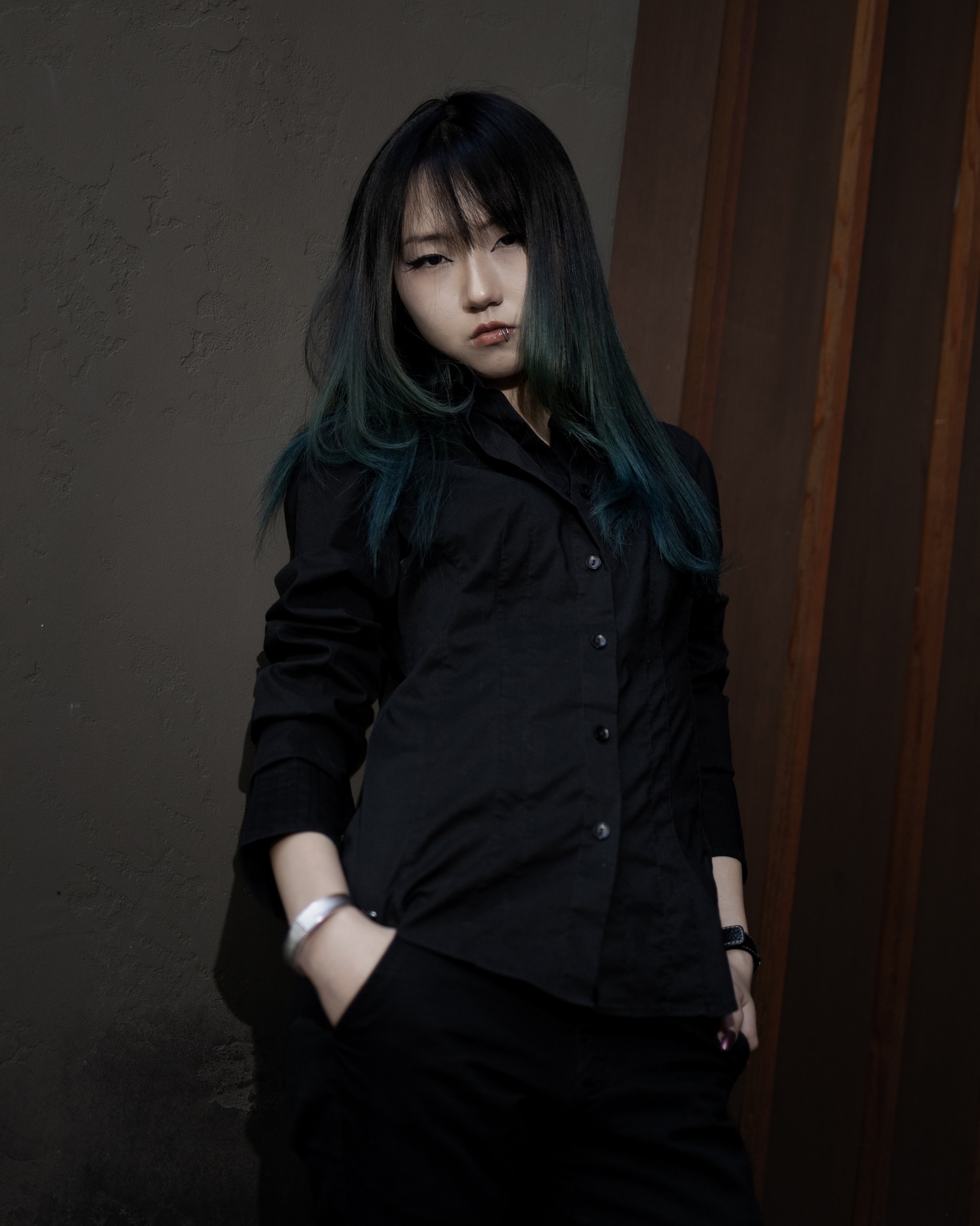 A young woman with long dark hair with green tips, wearing a black shirt and bracelets, standing against a dark textured wall and wooden panels, looking directly at the camera with a serious expression.