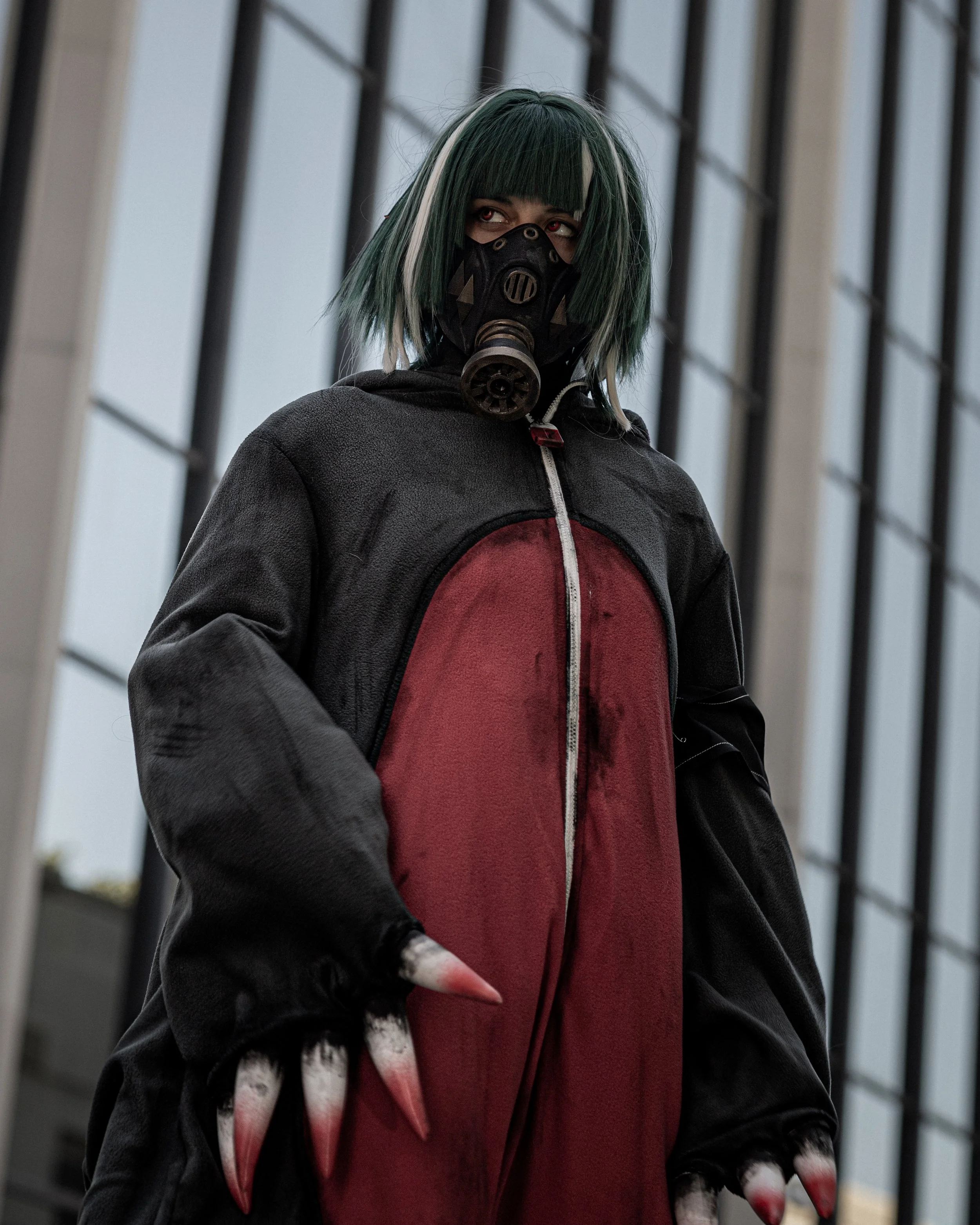 A person dressed as a character with green and white hair wearing a black gas mask and a dark hoodie over a red shirt, standing outdoors near a modern glass building.