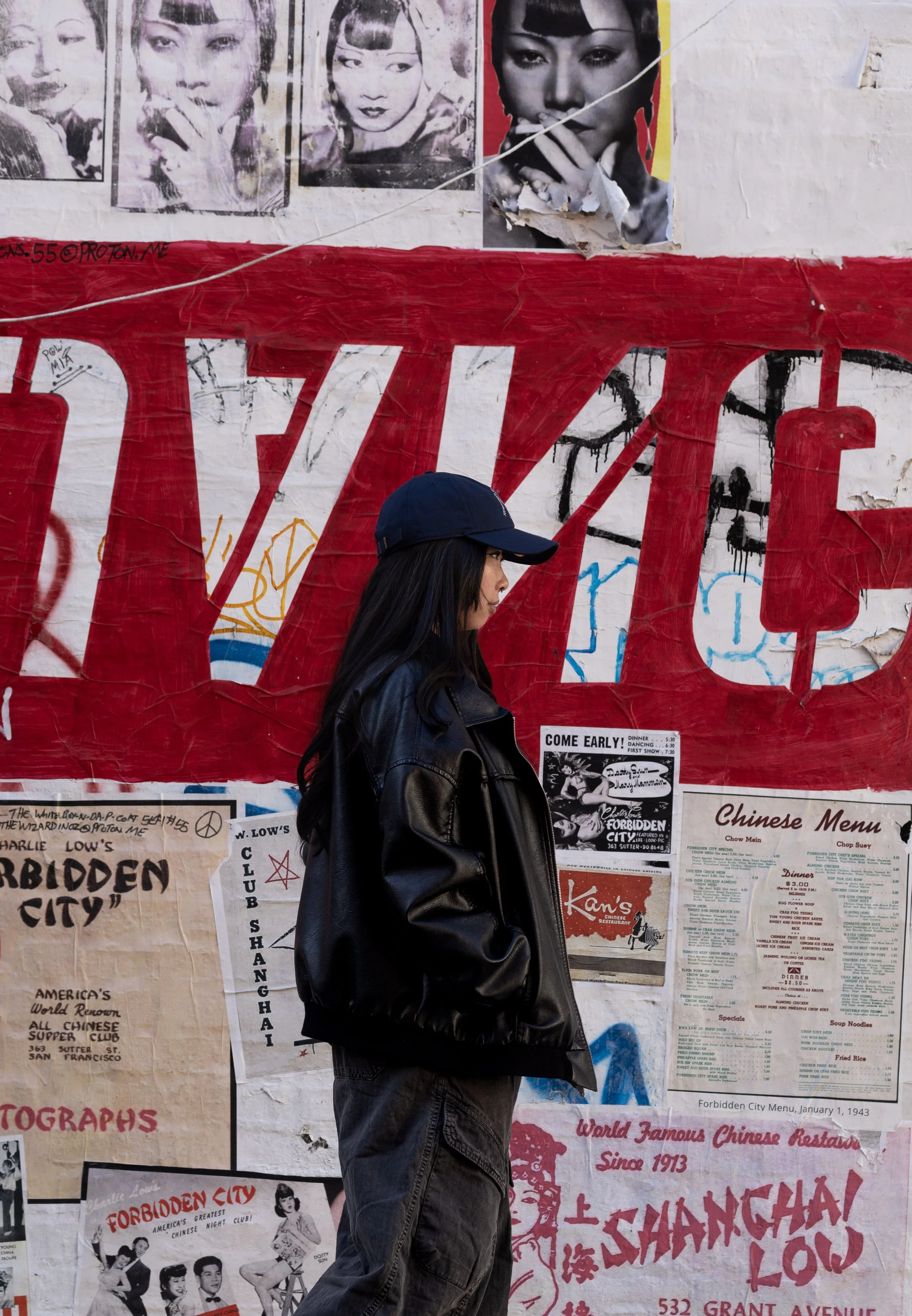 A person walking past a wall with posters, photographs, and advertisements, including black and white images of women and Chinese menu listings. The person is wearing a black cap and jacket, with long dark hair.