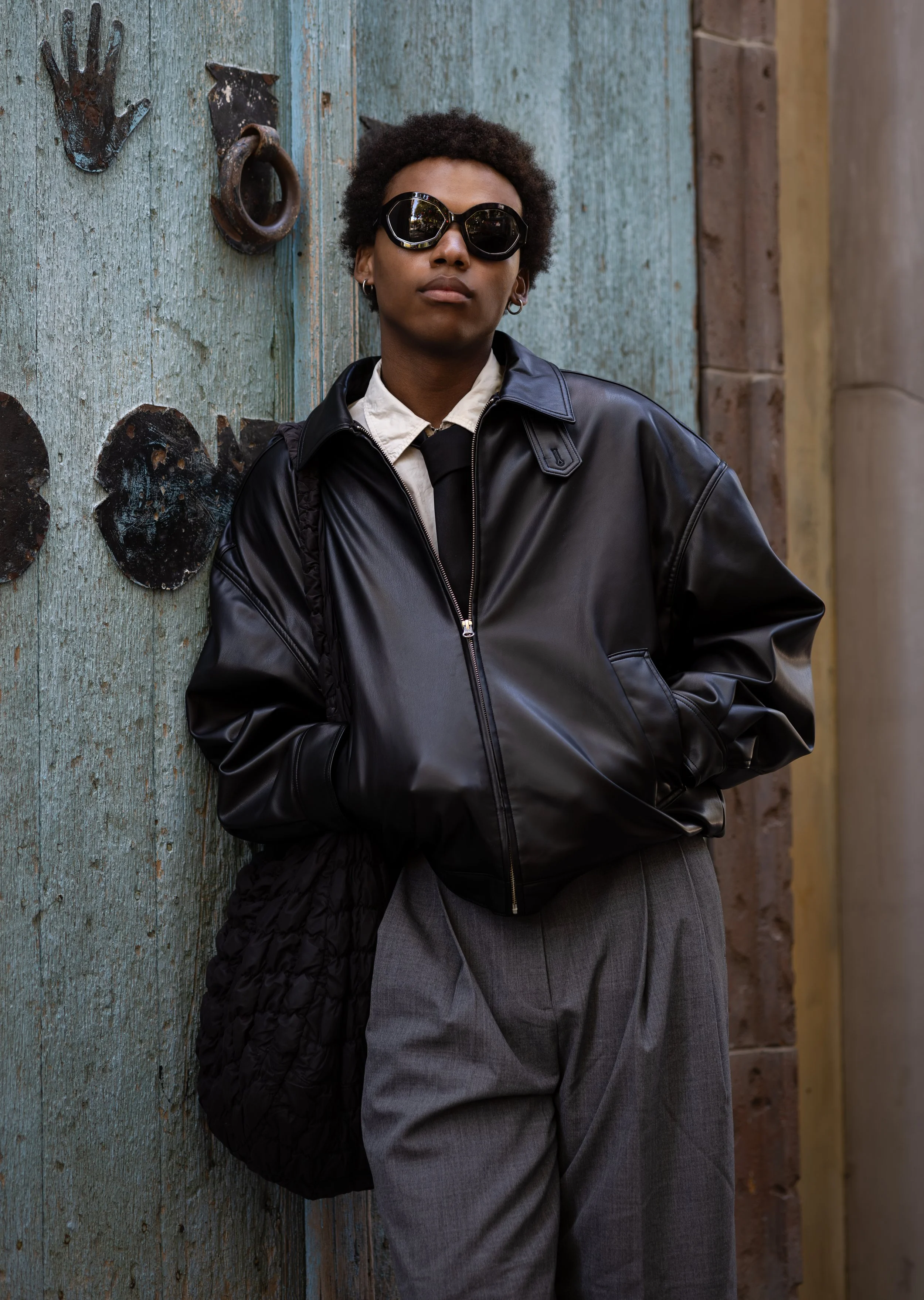 Person wearing black sunglasses, a black leather jacket, a white shirt, and grey pants, leaning against a weathered blue wooden wall with metal decorations.