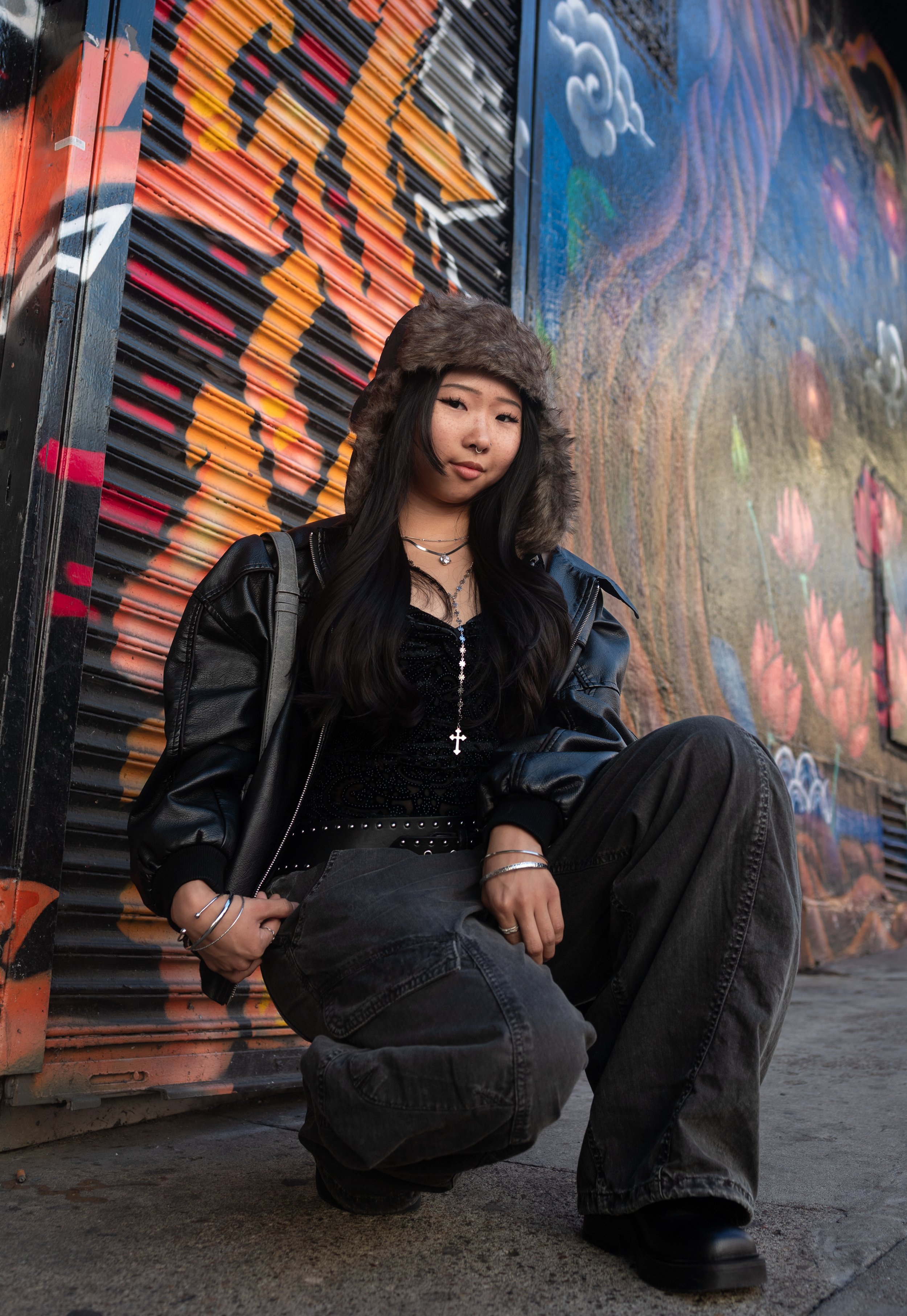 A young woman with long black hair, wearing a furry winter hat, black jacket, and gray jeans, sitting against a colorful graffiti wall.
