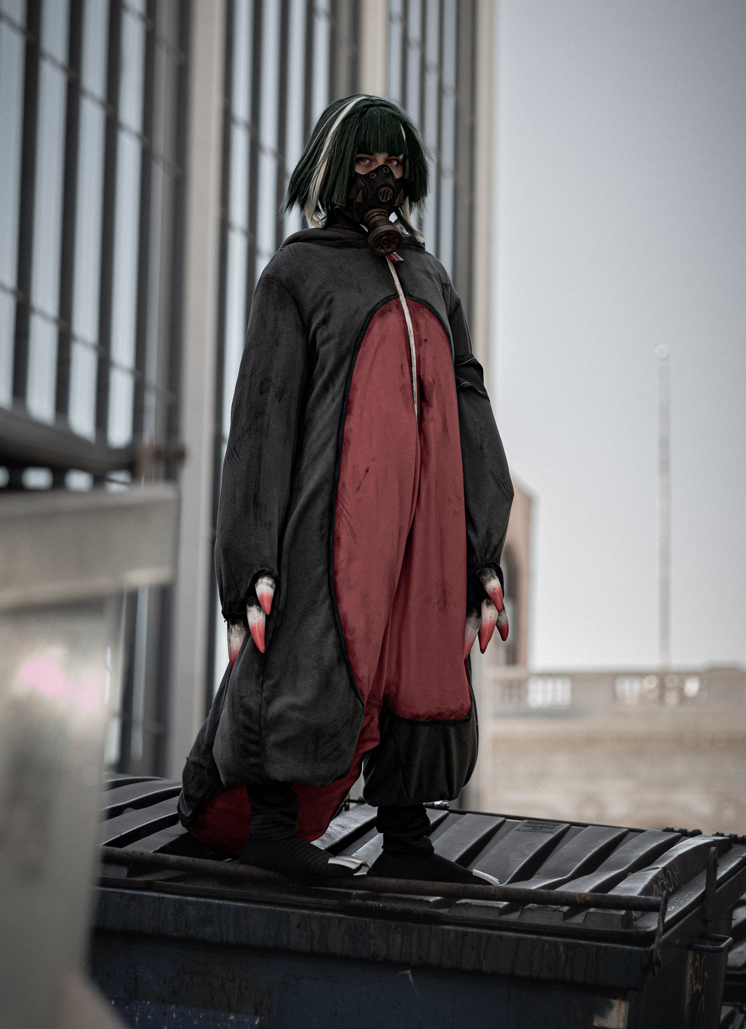 Person with green and black hair wearing a gas mask and black and red jacket sitting on a bench next to a trash bin outside near a building with large windows.