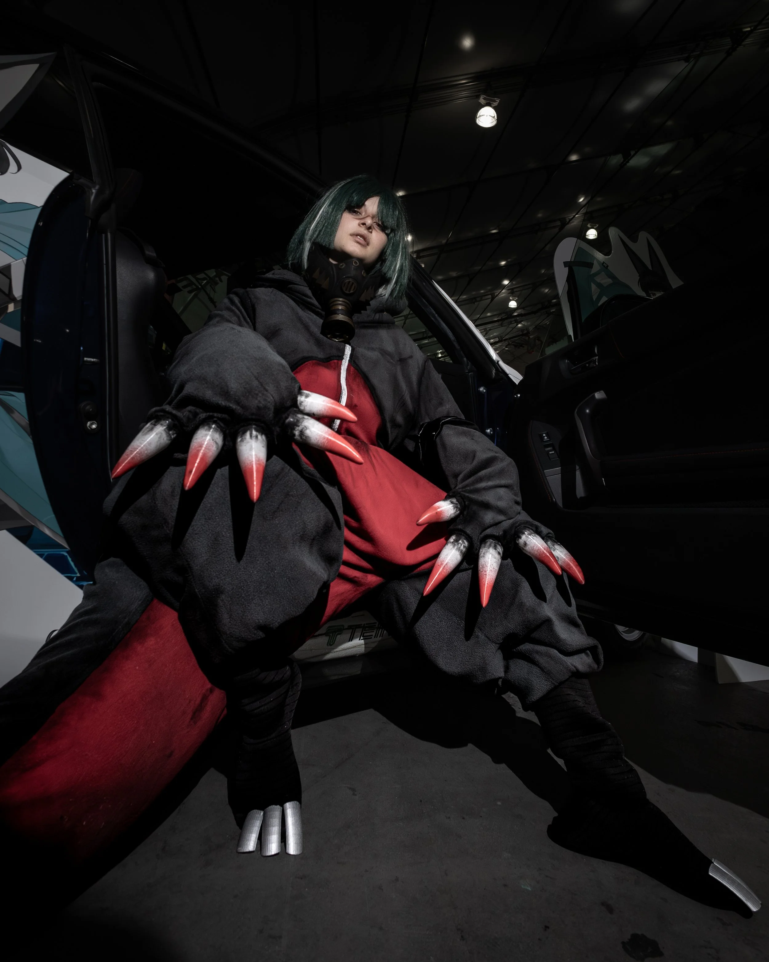 Person in costume resembling a character from a movie or video game, wearing a gas mask, black jacket, red accents, with claw-like gloves with long, pointed, metallic claws. They are sitting in a vehicle, possibly a race car, inside a large, dimly li