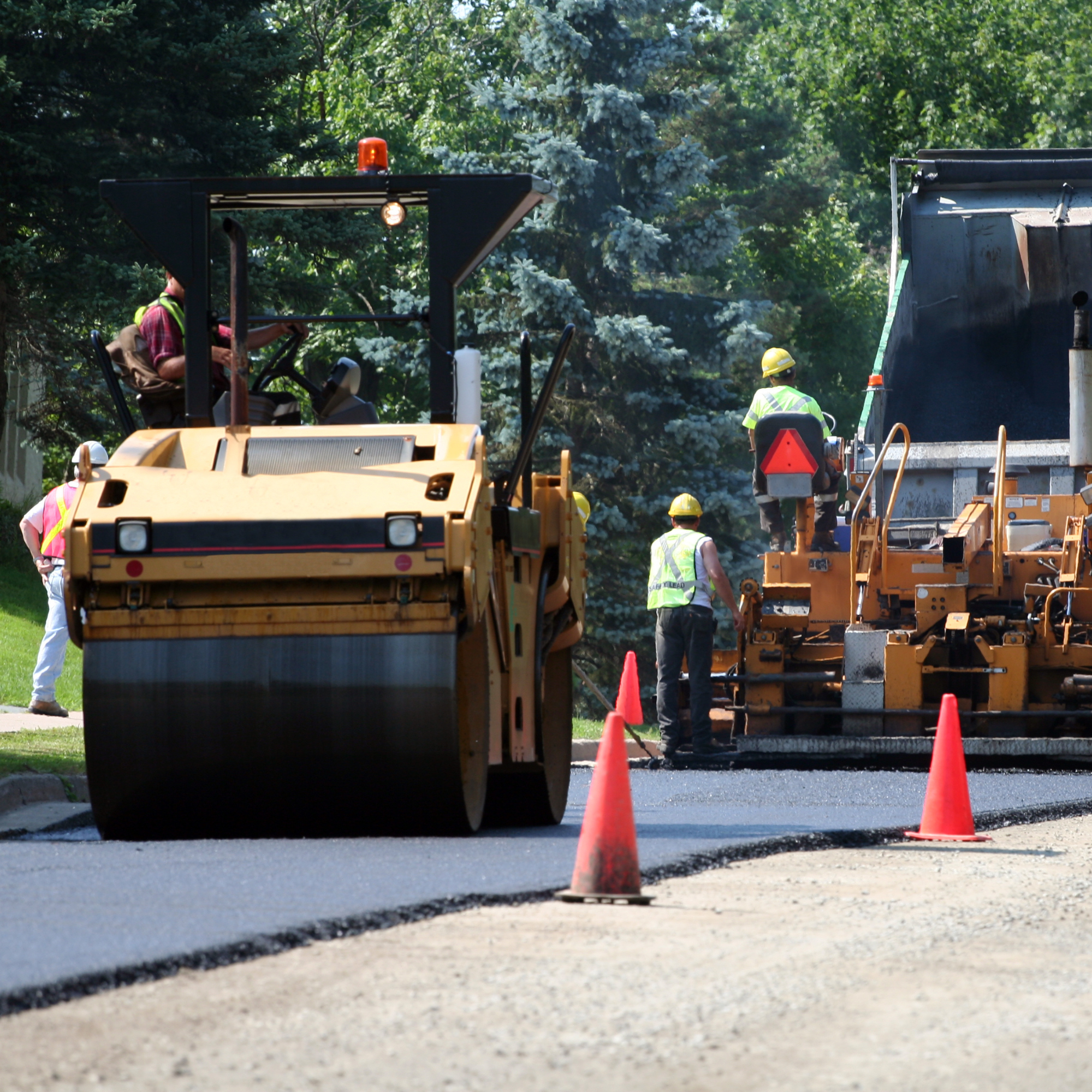 Workers paving asphalt on a road with construction equipment and orange traffic cones.