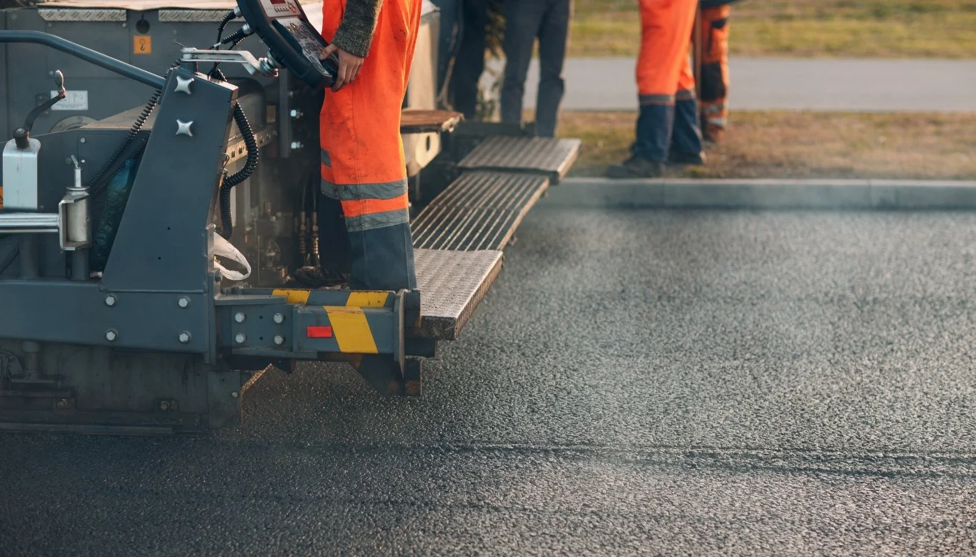 Workers in orange safety uniforms paving a road with asphalt.