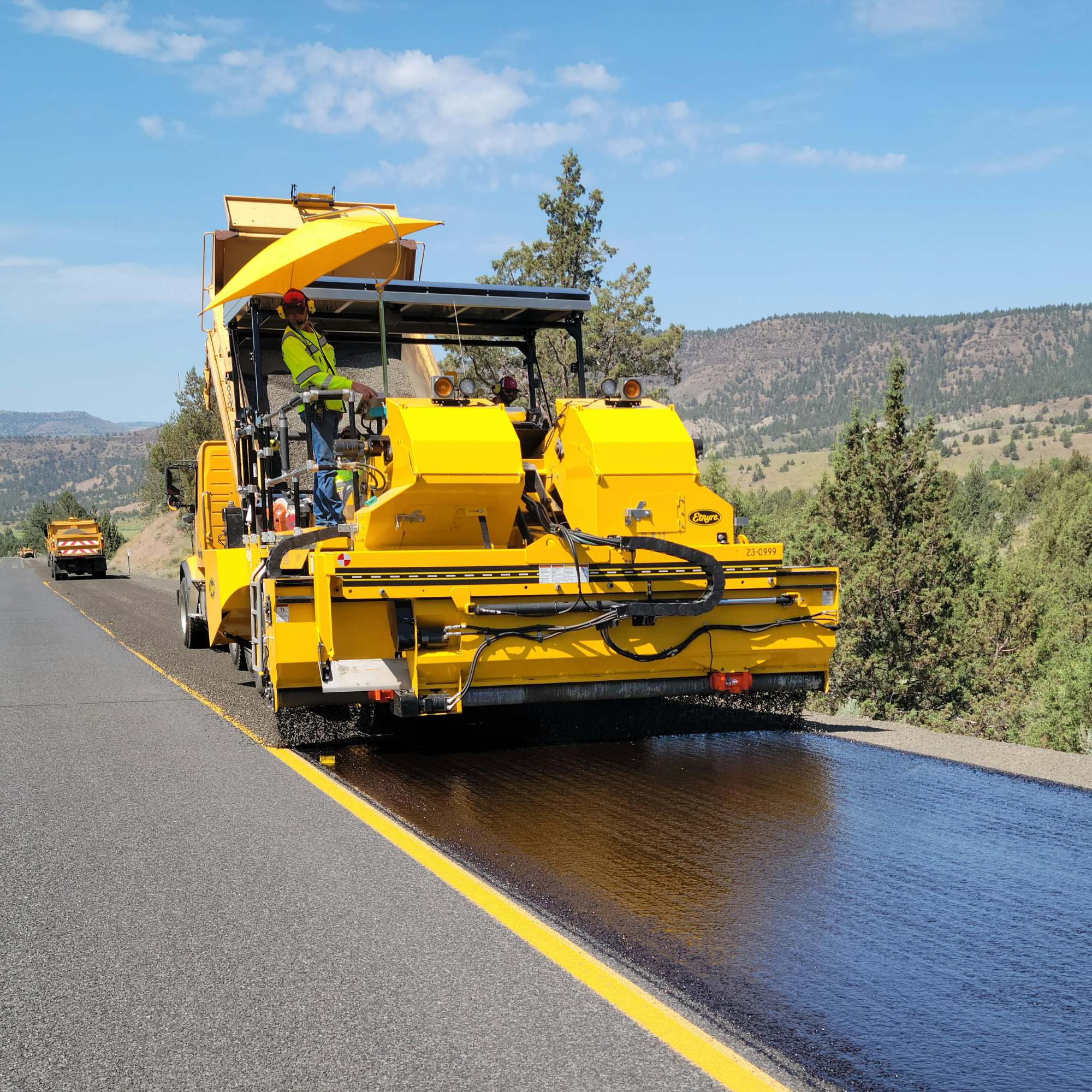 A yellow road construction vehicle spreading hot asphalt on a multi-lane road with mountains and blue sky in the background.
