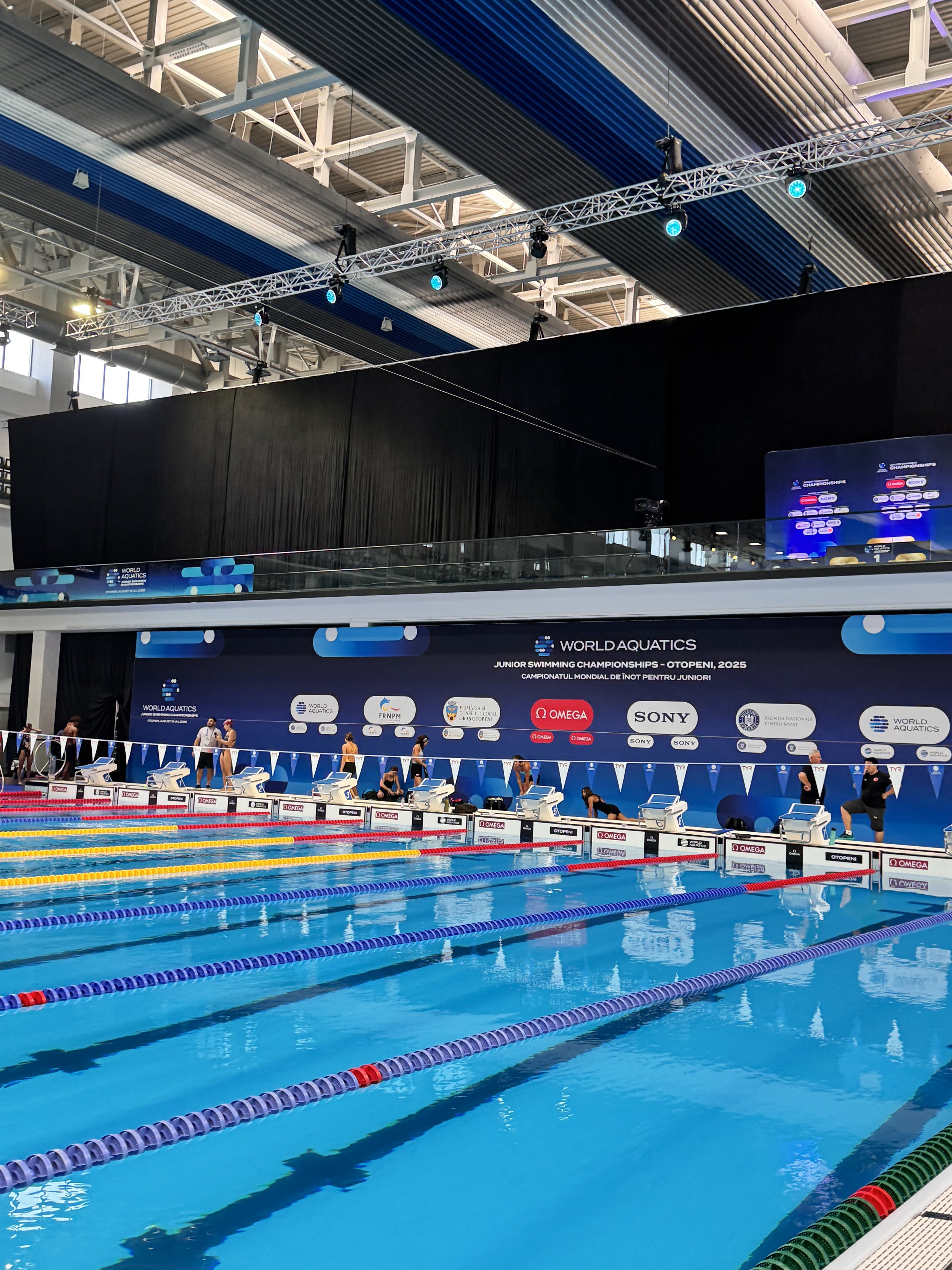 An indoor swimming pool at a competitive swimming event with multiple lanes, a large digital scoreboard, and a black backdrop displaying event information. There are swimmers and officials preparing at the edge of the pool.