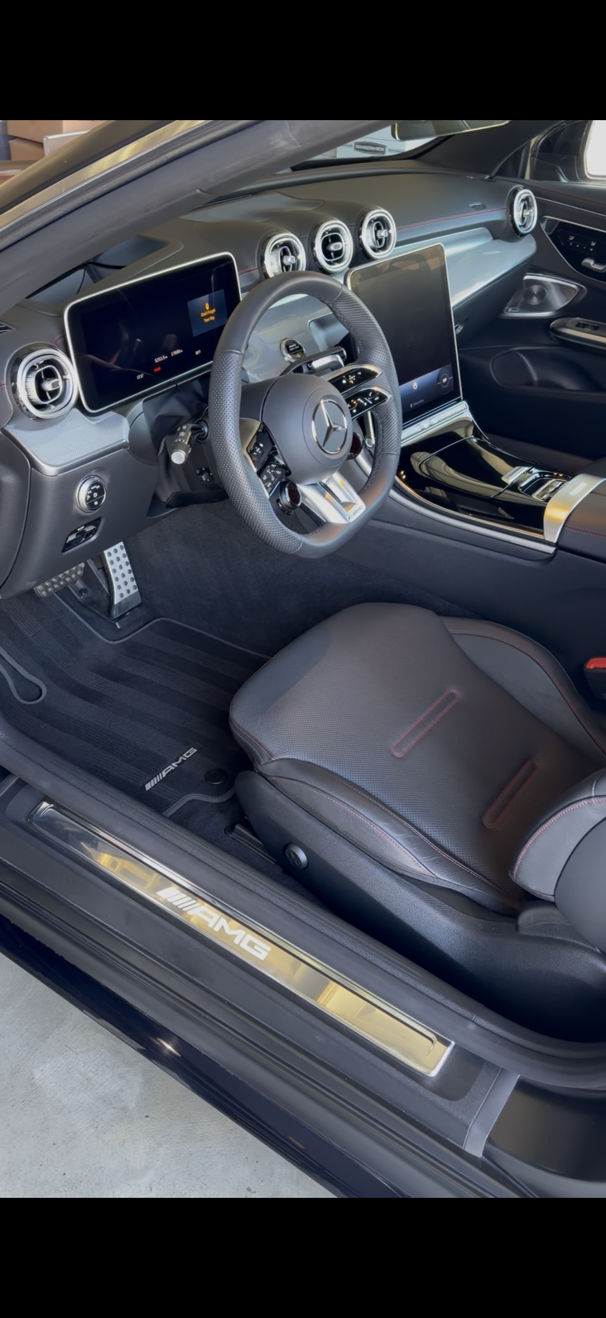 Interior of a modern luxury Mercedes-Benz car, showing a leather seat, steering wheel, digital displays, air vents, and AMG branding on the door sill.
