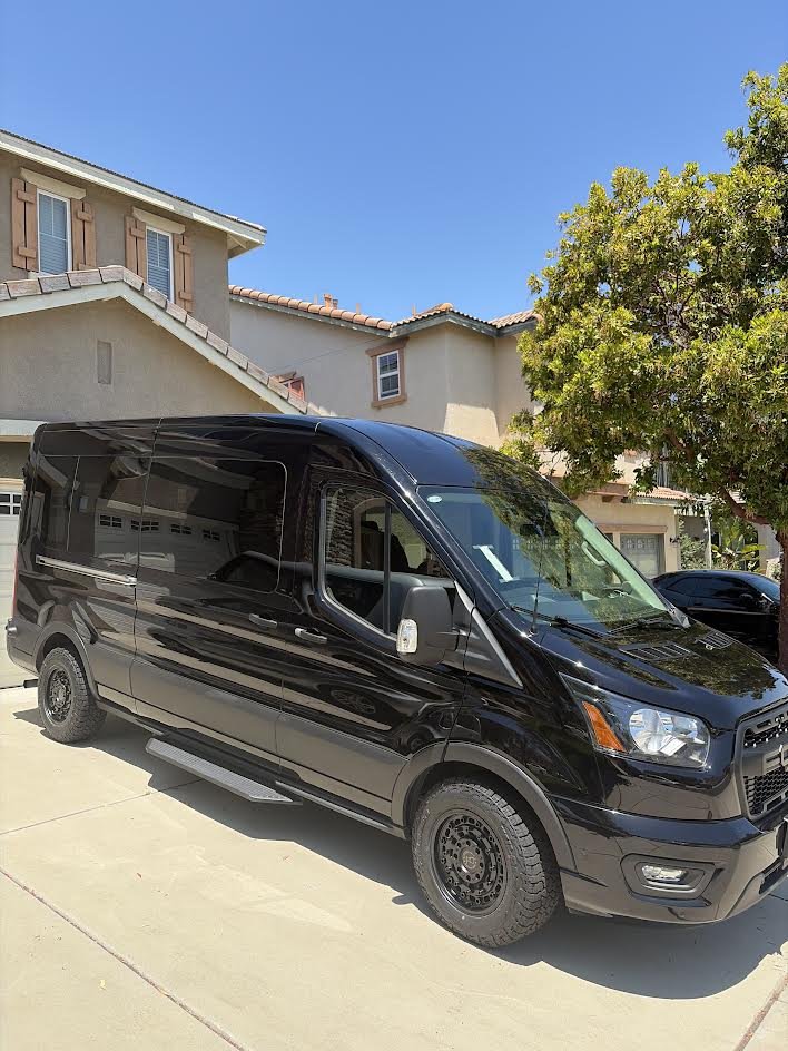 Black cargo van parked in a suburban driveway on a sunny day, with a beige house, a green tree, and a clear blue sky in the background.