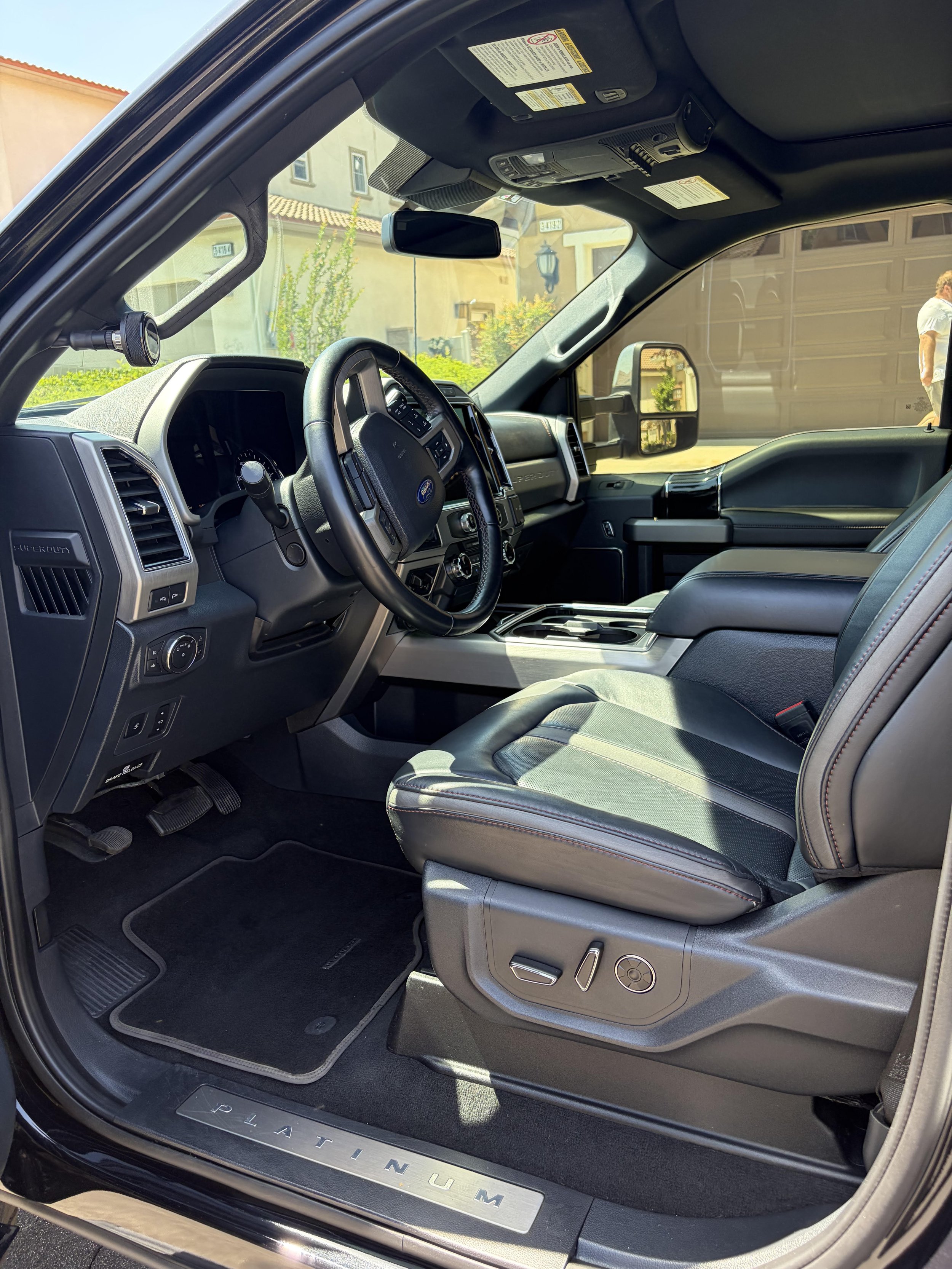 The image shows the interior of a Ford pickup truck. The view is from the driver's side door, showing the dashboard, steering wheel, center console, and front passenger seat. The truck has a black interior with leather seats, and the dashboard features various controls. The door sill has a metal plate with the text 'PLATINUM UM.' The background outside the truck includes buildings and a person walking.
