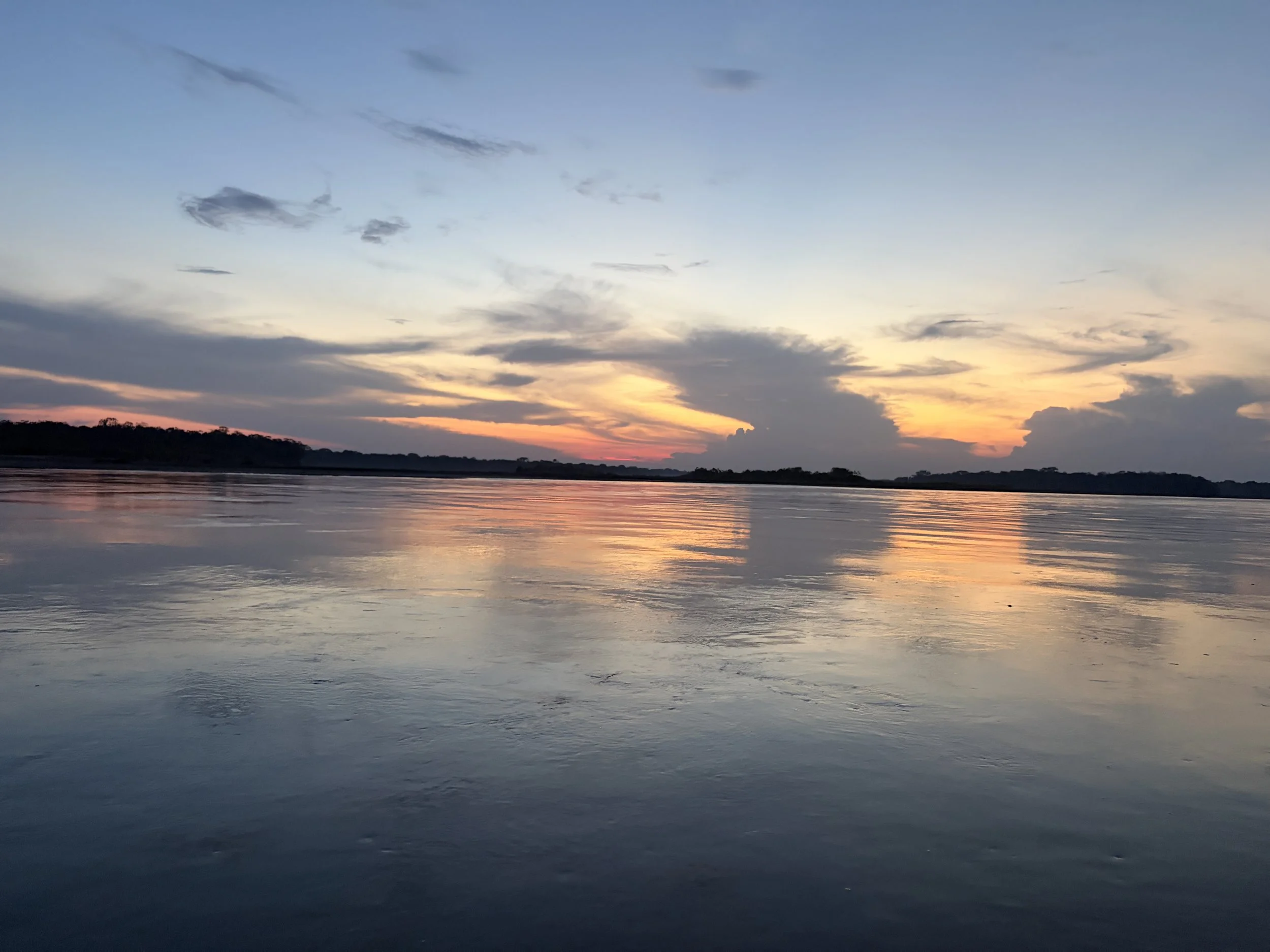 A sunset over a calm river with a partly cloudy sky and a forested shoreline in the distance.