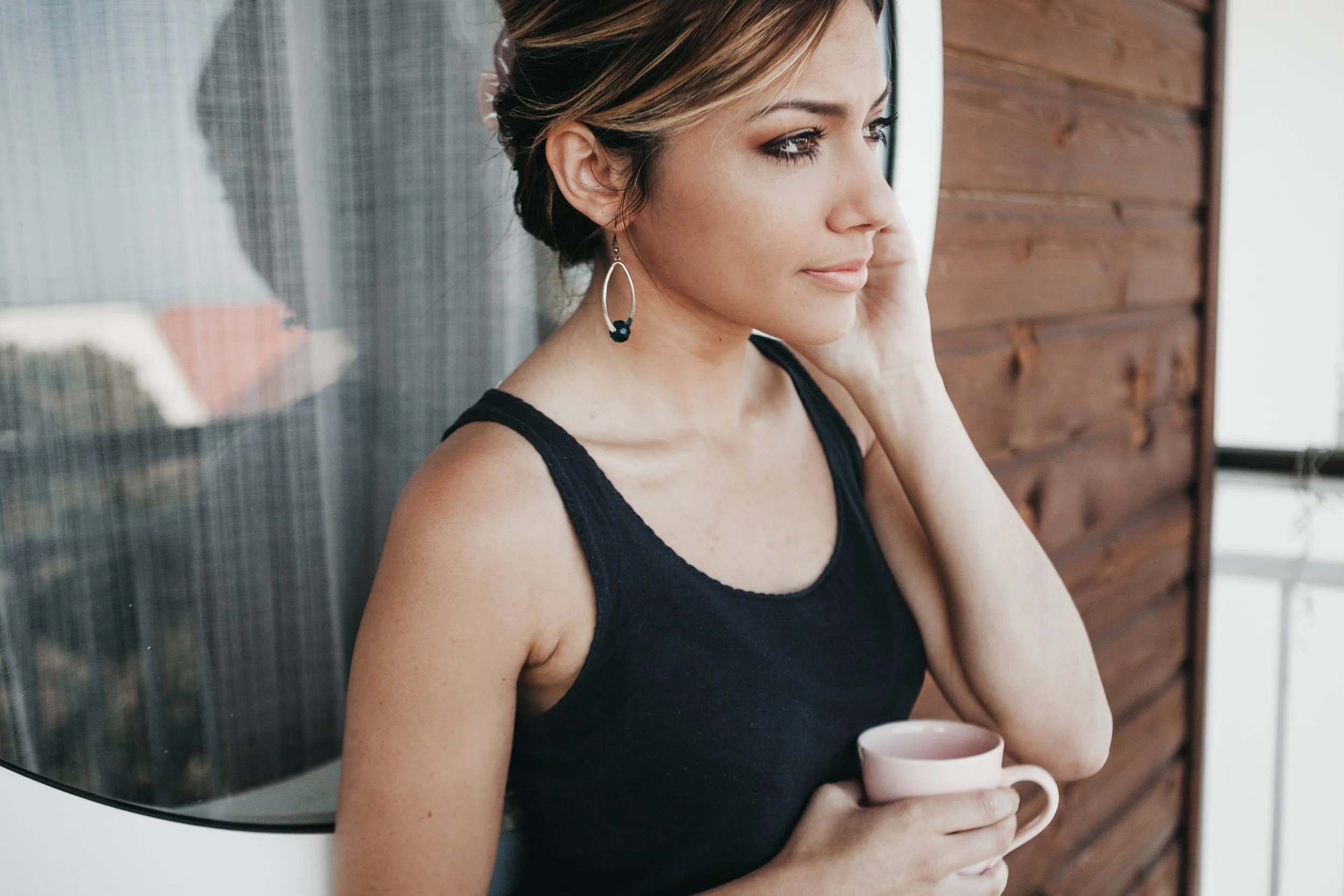 A woman with short hair, wearing earrings and a black sleeveless top, holding a white mug while standing near a wooden wall.