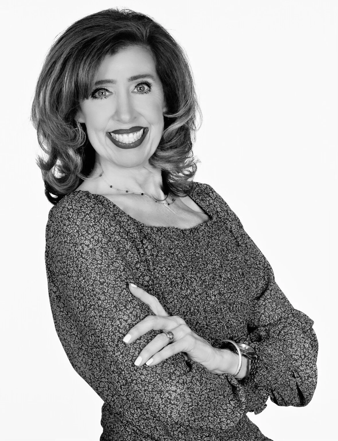 Black and white portrait of a woman with shoulder-length hair, smiling, wearing a patterned blouse, with her arms crossed.