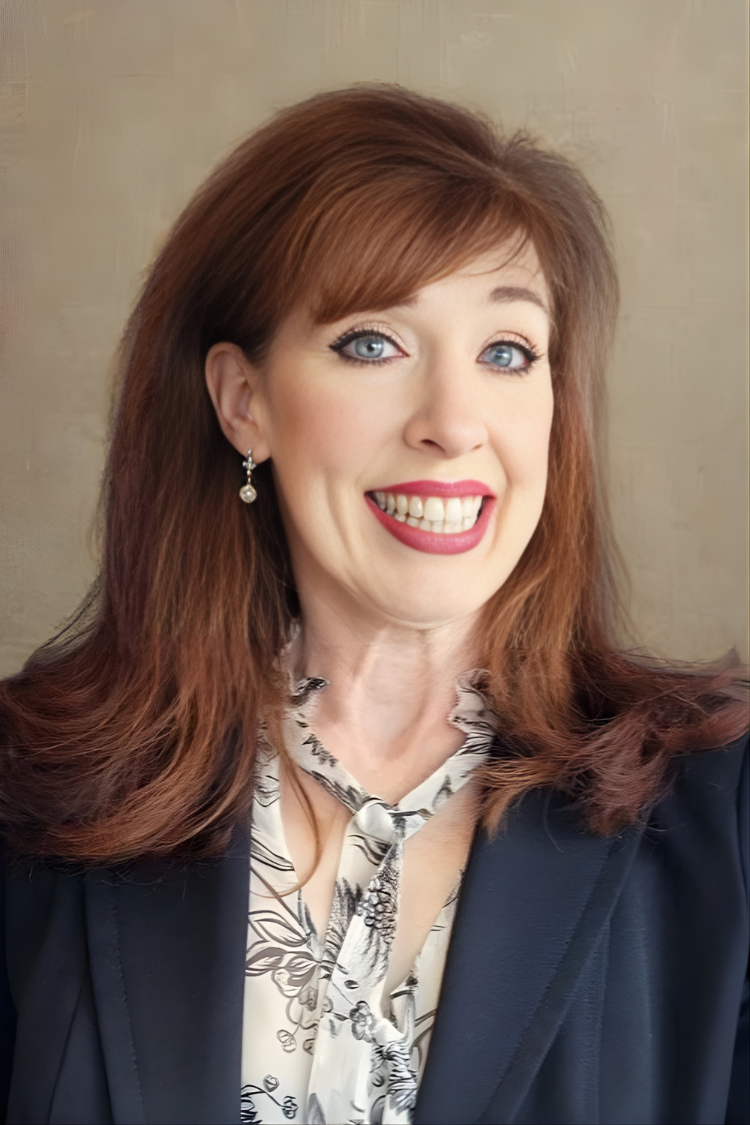 A woman with shoulder-length reddish-brown hair, light blue eyes, and wearing earrings, a patterned blouse, and a dark blazer, smiling at the camera.