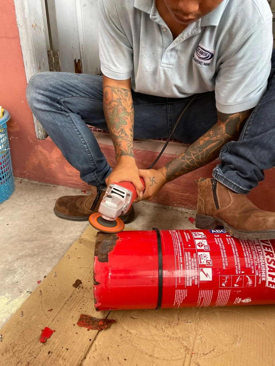 A man with tattoos wearing a gray polo shirt and jeans is using a handheld angle grinder to cut a red fire extinguisher on a cardboard surface. His feet are apart, and he is wearing brown work boots.