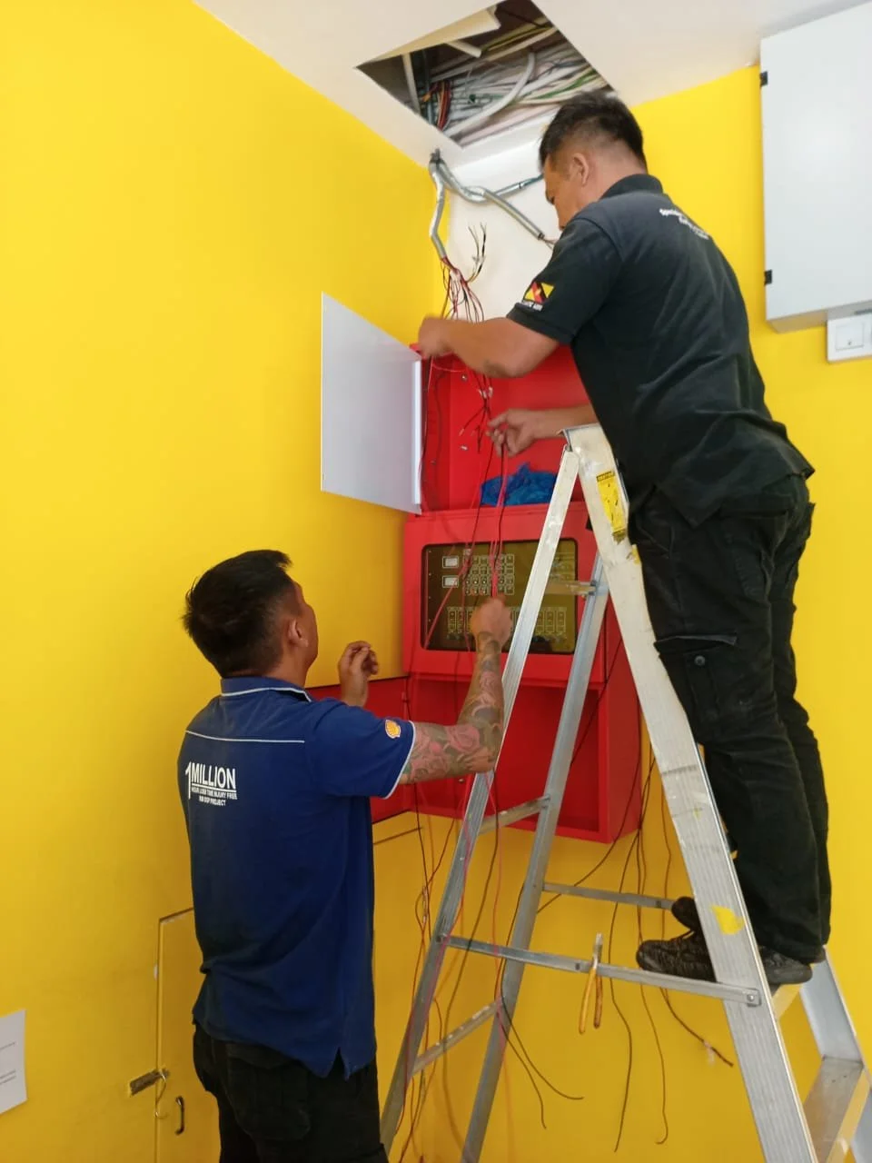 Two electricians working on wiring inside an electrical panel with an open ceiling hatch in a room with yellow walls.