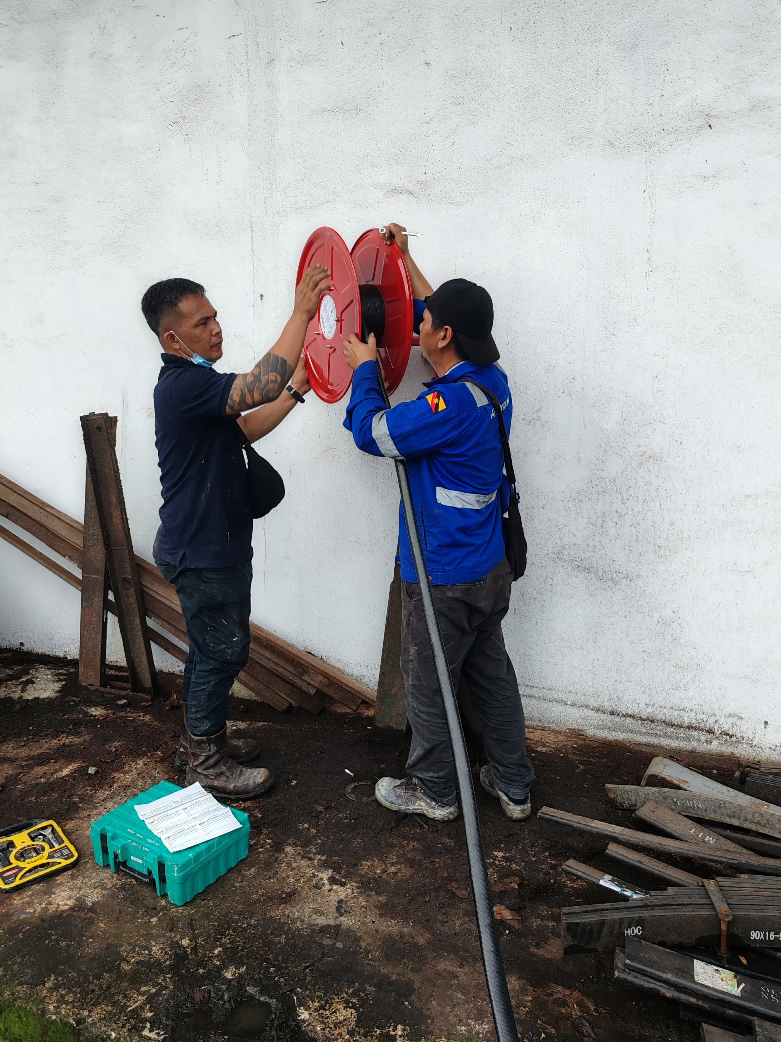 Two men working together on a task involving a red fire hose reel mounted on a white wall, one holding the reel and the other using a tool. They are outdoors on muddy ground with tools and equipment scattered around.