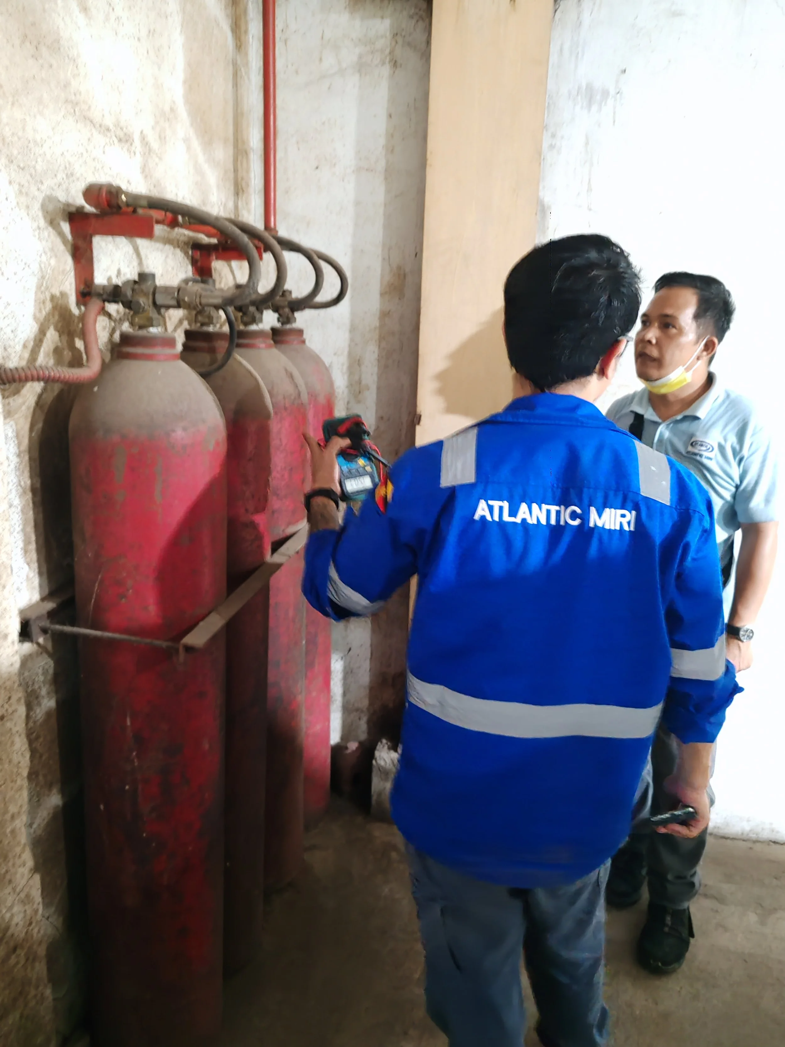 Two men are standing near rusted fire extinguishers mounted on a concrete wall. One man, wearing a blue jacket labeled "Atlantic Miri," is holding a device, while the other, wearing a white shirt and face mask, is looking at him.
