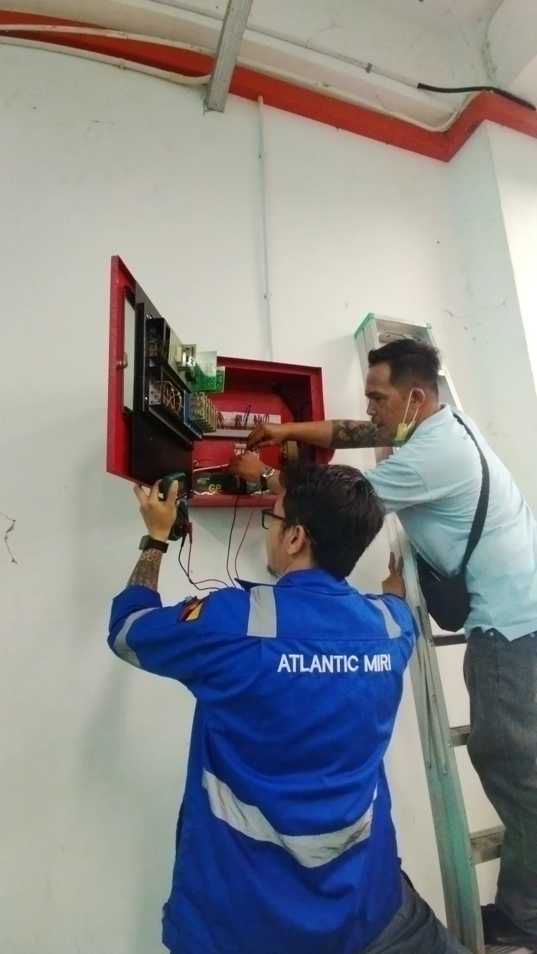 Two technicians installing or repairing electrical equipment in a red metal cabinet on a white wall, one standing on a ladder and the other kneeling.