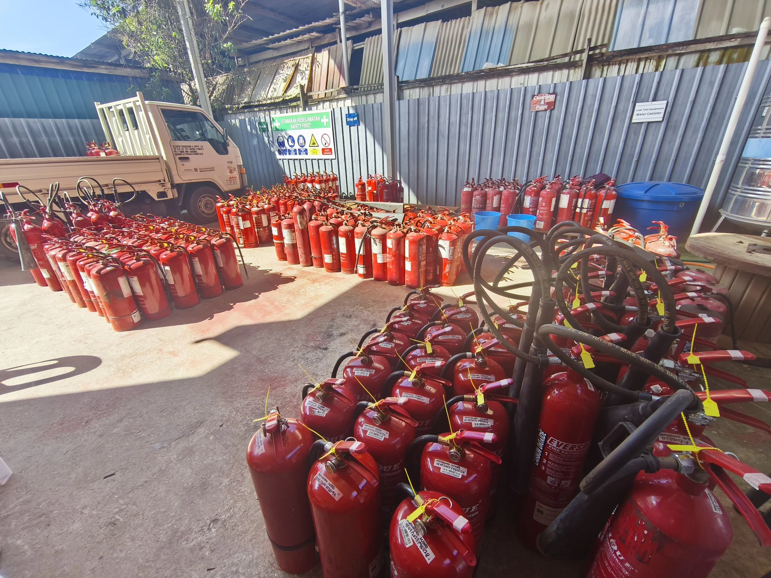 An outdoor storage area filled with numerous red fire extinguishers, some on the ground and some mounted on carts. A small truck is parked to the left, and there are signs about safety and equipment on the corrugated metal fence behind.