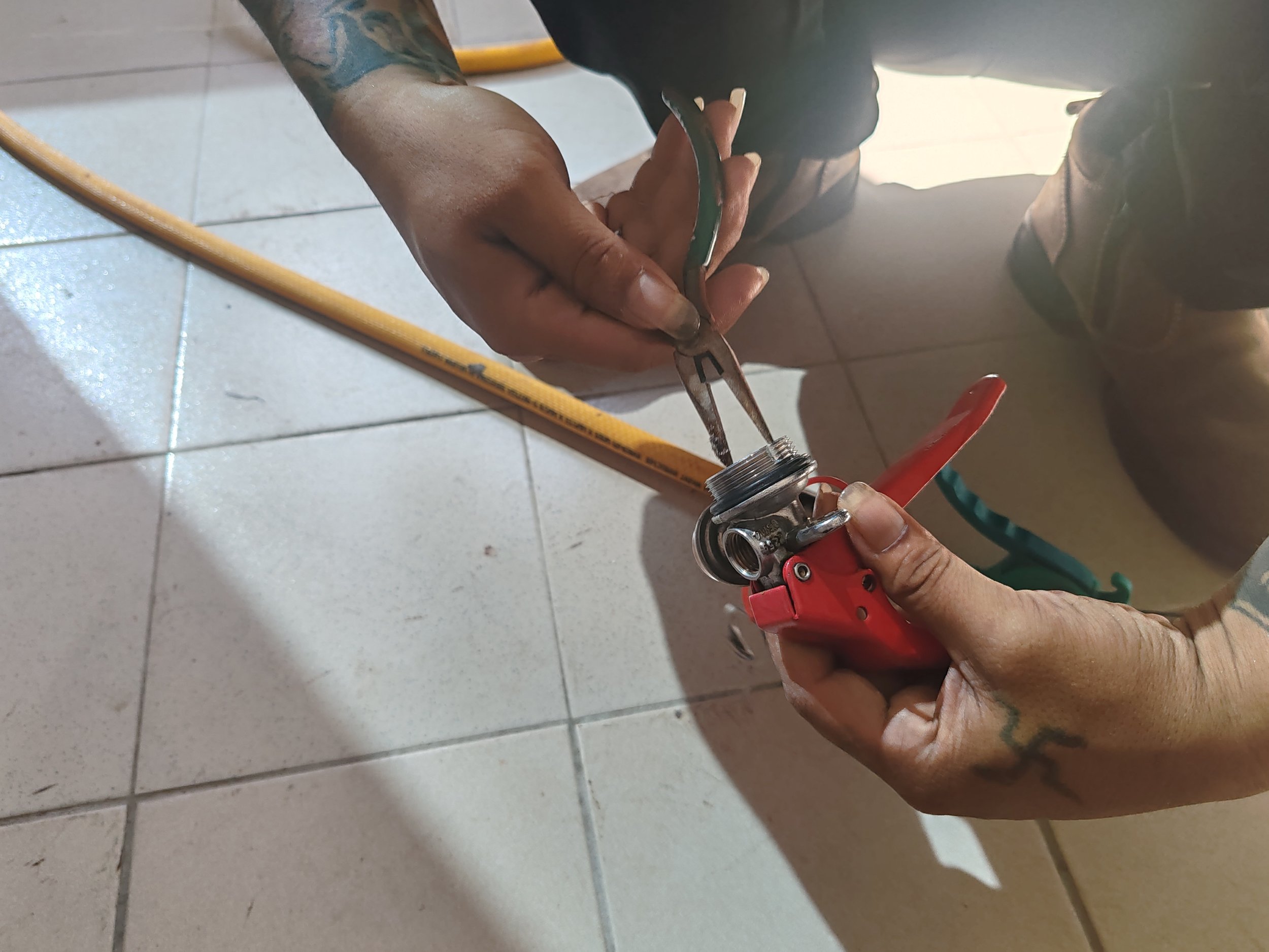 A person with tattoos on their arm is working on a plumbing fixture, holding a wrench and adjusting a metal pipe fitting on a tiled floor, with a yellow pipe and red-handled tool nearby.