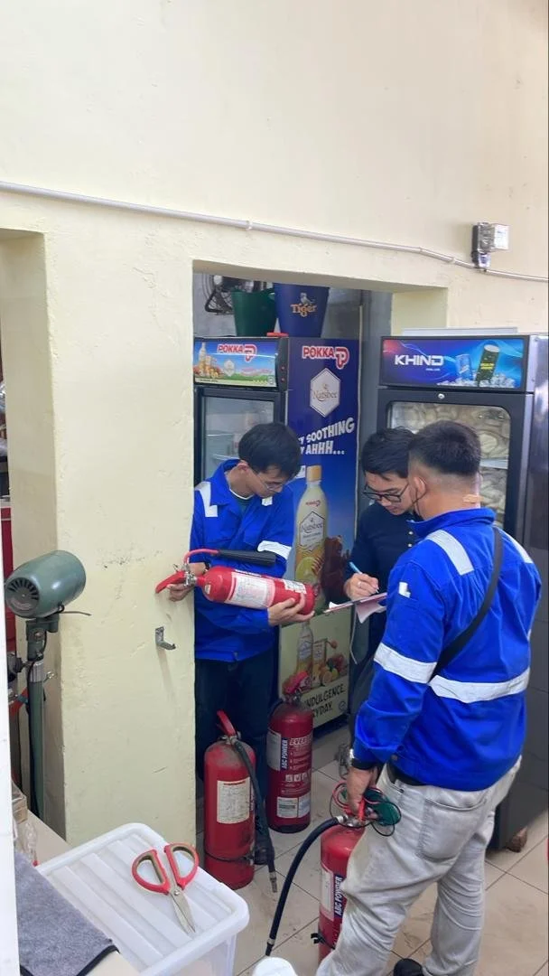 Three men in blue jackets are inspecting fire extinguishers near vending machines in a small room.