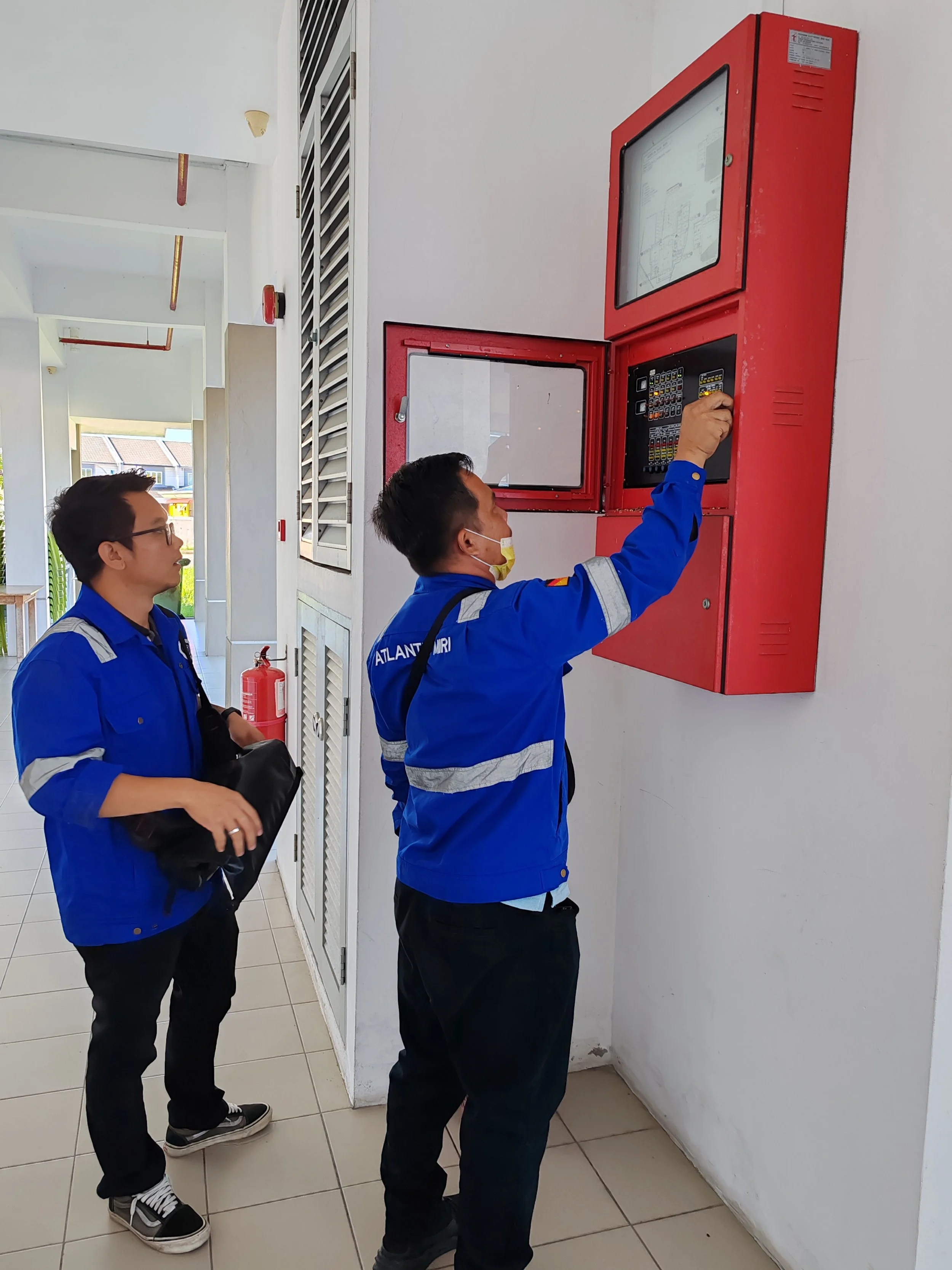 Two men in blue uniforms are servicing a red fire alarm control panel on a white wall. One man is reaching for the controls, while the other observes holding a black bag. Fire extinguisher is visible on the wall nearby.