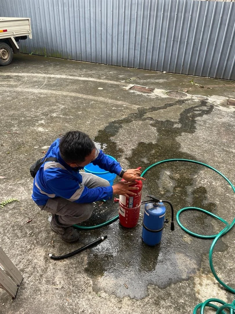 Man in blue jacket crouching on wet concrete, pressing a fire extinguisher. A blue and black air compressor and green hoses are nearby. Metal siding wall and a small truck are in the background.