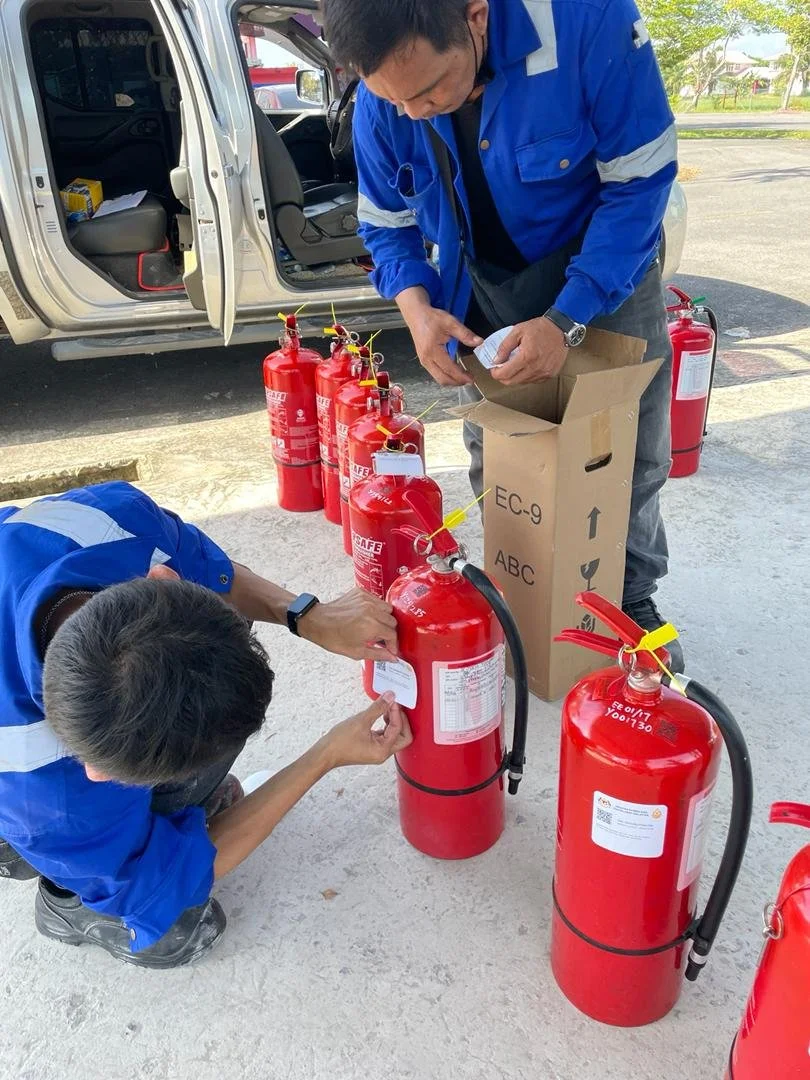 Two workers are preparing or inspecting fire extinguishers outdoors, with several fire extinguishers lined up on the ground and one in a box, near a vehicle with its door open.