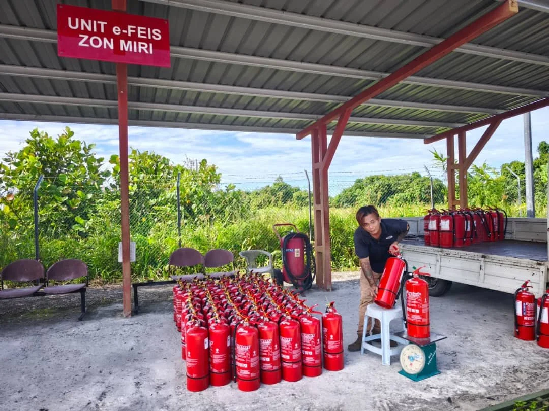 A man arranging red fire extinguishers under a shelter labeled 'UNIT e-FEIS ZON MIRI,' with more fire extinguishers on a truck bed nearby. There are three chairs and greenery background.