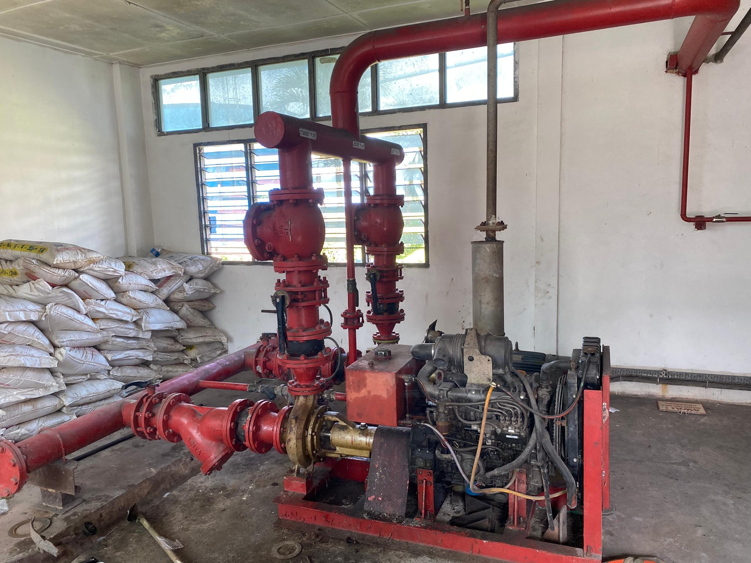 Red fire pump system with pipes and valves in a room under construction. Window with bars and stacking bags are visible in the background.