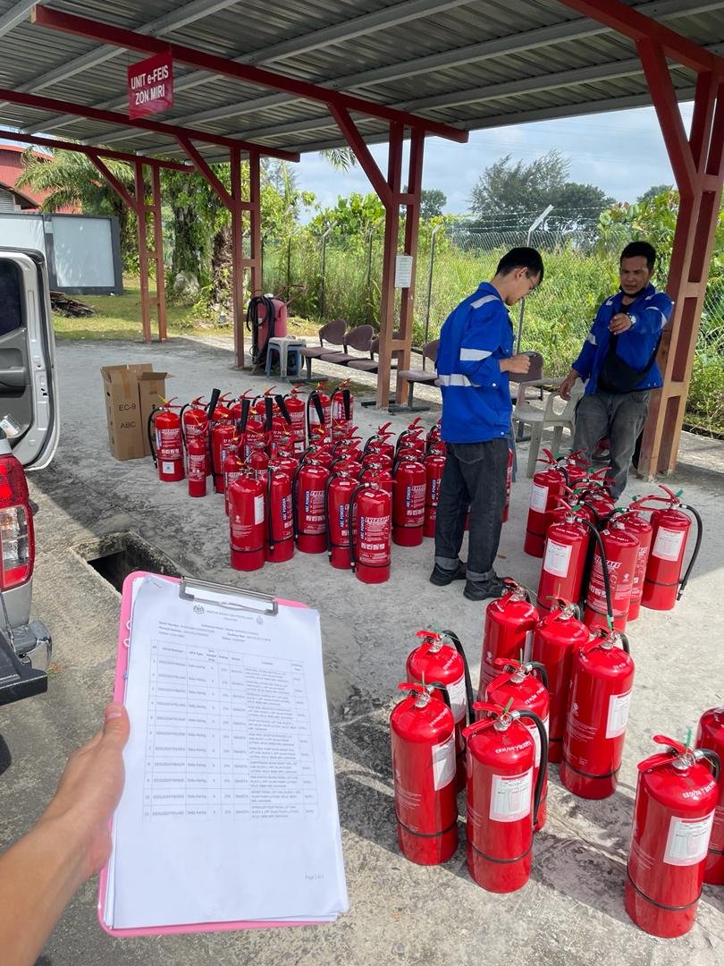 A person holding a clipboard with a list stands near multiple fire extinguishers arranged on the ground under a shelter. Two individuals in blue uniforms are inspecting the fire extinguishers.