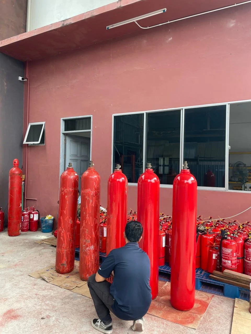 A man crouching in front of several large red fire extinguishers outside a building with a red wall and large windows.