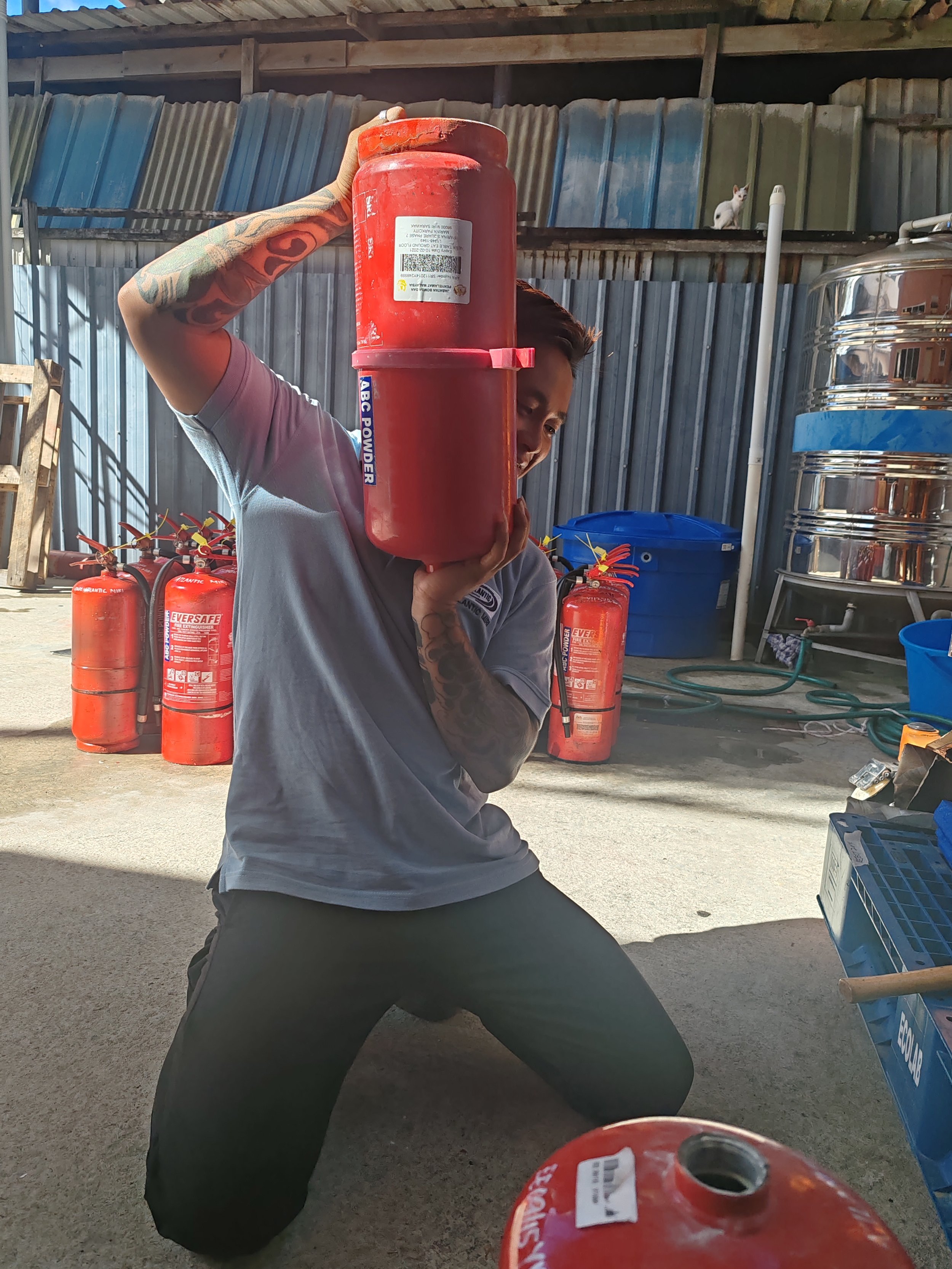 A person in a kneeling position holding a red fire extinguisher on their shoulder while smiling. Several other fire extinguishers are visible in the background against a corrugated metal wall, along with a blue recycling bin, some hoses, and a small 