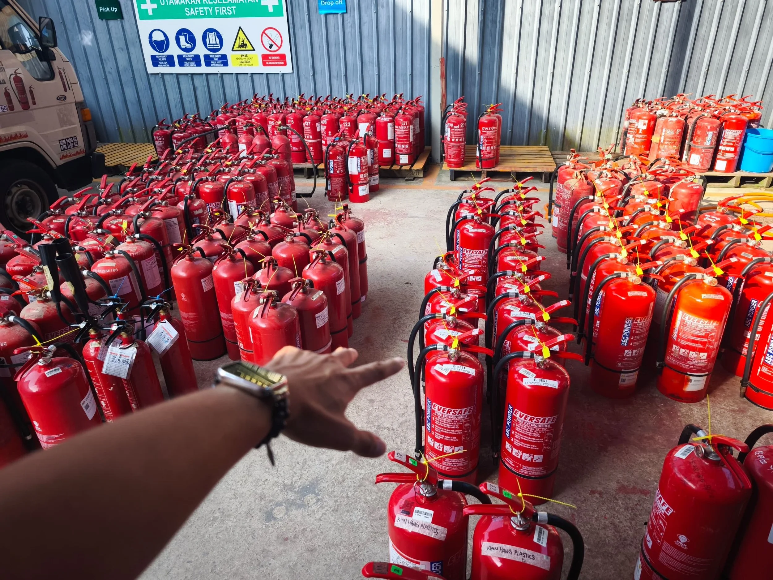 Multiple red fire extinguishers arranged in rows on the ground and on wooden pallets at an outdoor storage area, with safety signs on a blue corrugated metal wall behind them.