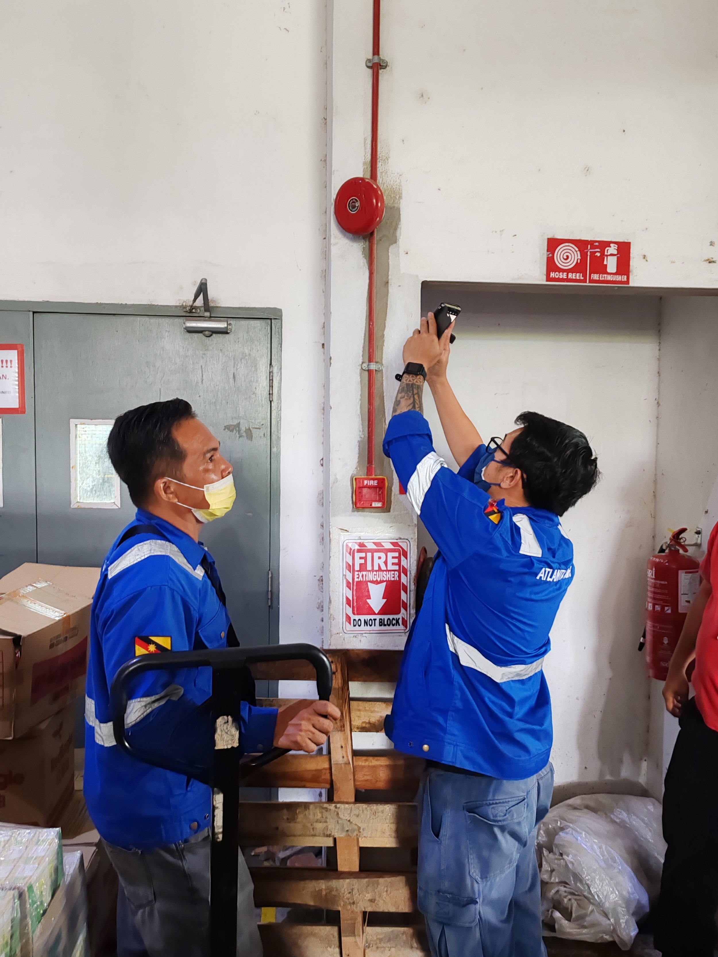 Two men in blue uniforms, one wearing a yellow face mask, are inspecting or installing a fire alarm and a fire safety system on a wall in an industrial or storage area. One man is holding a tool, and the other is holding a device or clipboard. There is a fire extinguisher to the right, and various boxes and bags are seen around them.