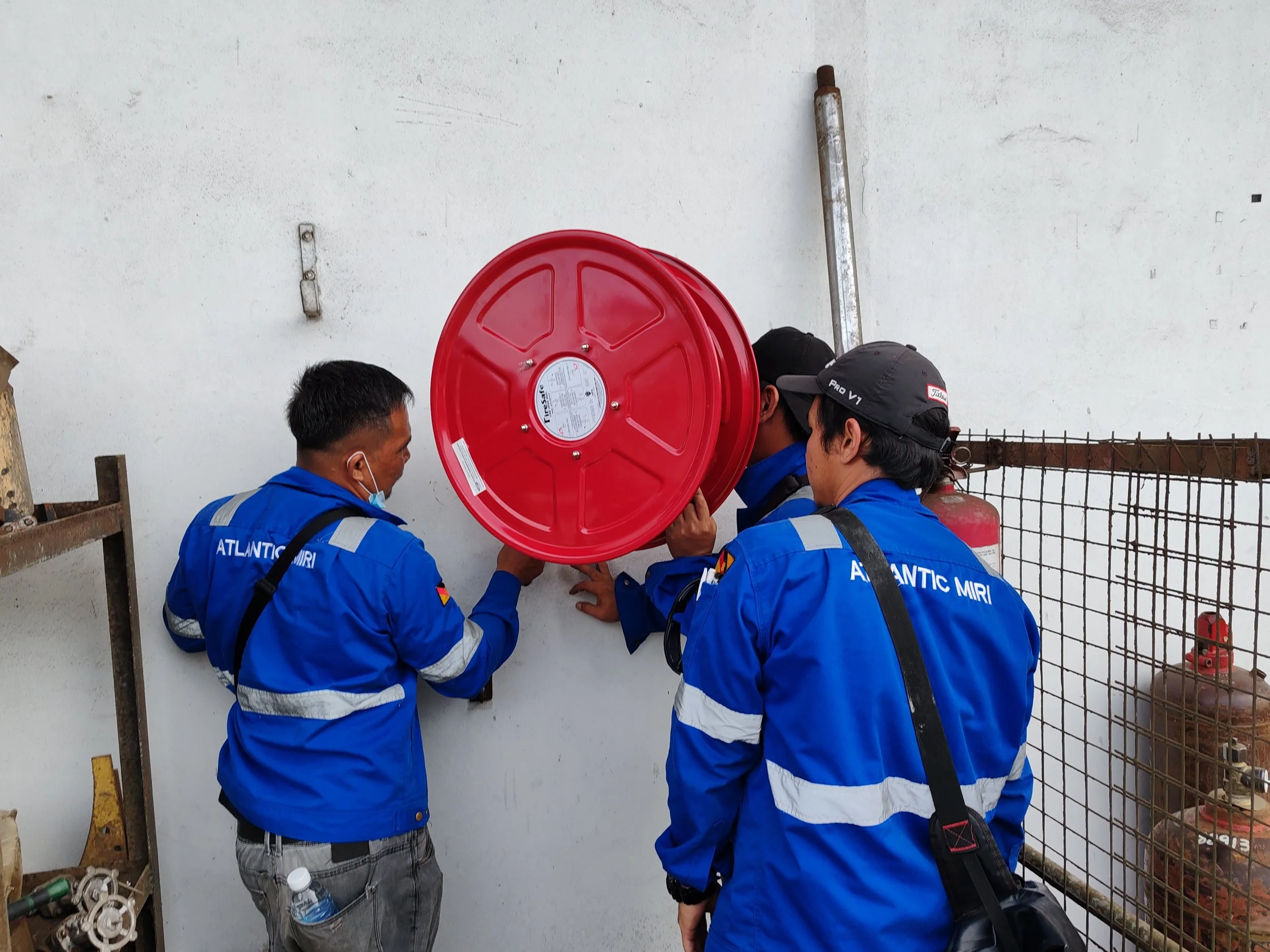 Three men in blue uniforms working together to install a fire hose reel on a white wall. One man is wearing a black cap and the others are wearing a face mask and no hat.