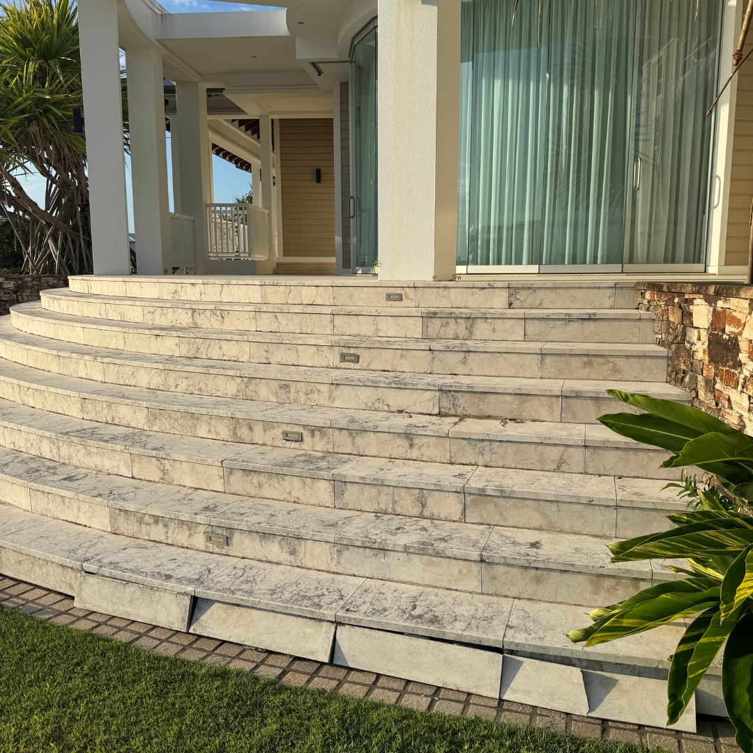 Exterior view of a house with curved stone steps leading to a glass sliding door. The house has light-colored siding and large windows with curtains, with some greenery and plants nearby.