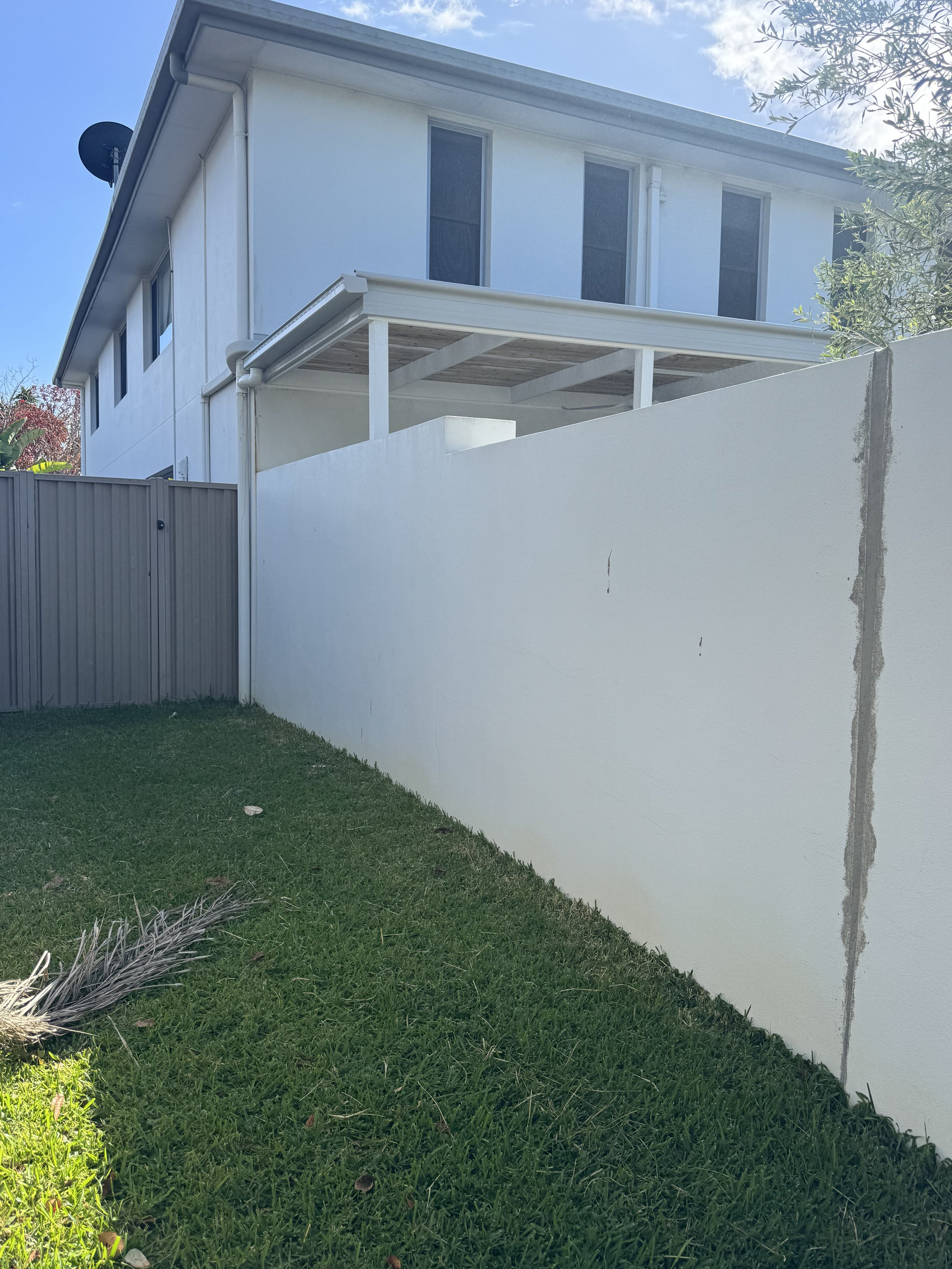 A two-story white house with a balcony and three tall, narrow windows on the upper floor. A satellite dish is attached to the corner of the house. The house has a section with a covered porch supported by white posts. In the foreground, there is a white wall and a grassy yard with a small dry palm branch. The sky is partly cloudy.