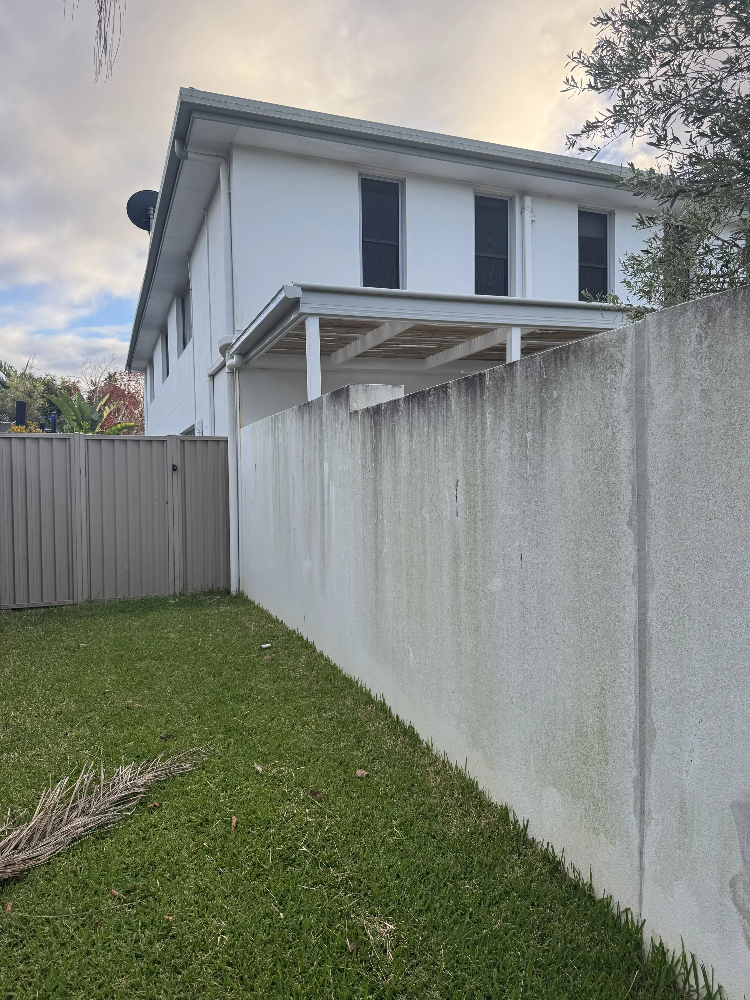 A white two-story house with three windows on the upper floor and an unfinished balcony or porch with white support beams. The house is surrounded by a concrete wall to the right and a gray metal fence on the left. The lawn in the foreground is green with a few small plants and some debris. The sky above is cloudy with patches of blue.