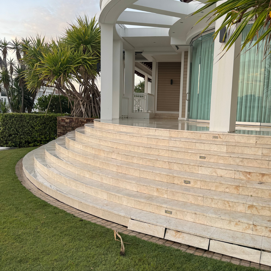 Exterior view of a modern house with beige marble curved stairs leading up to a porch, surrounded by green grass and tropical plants.