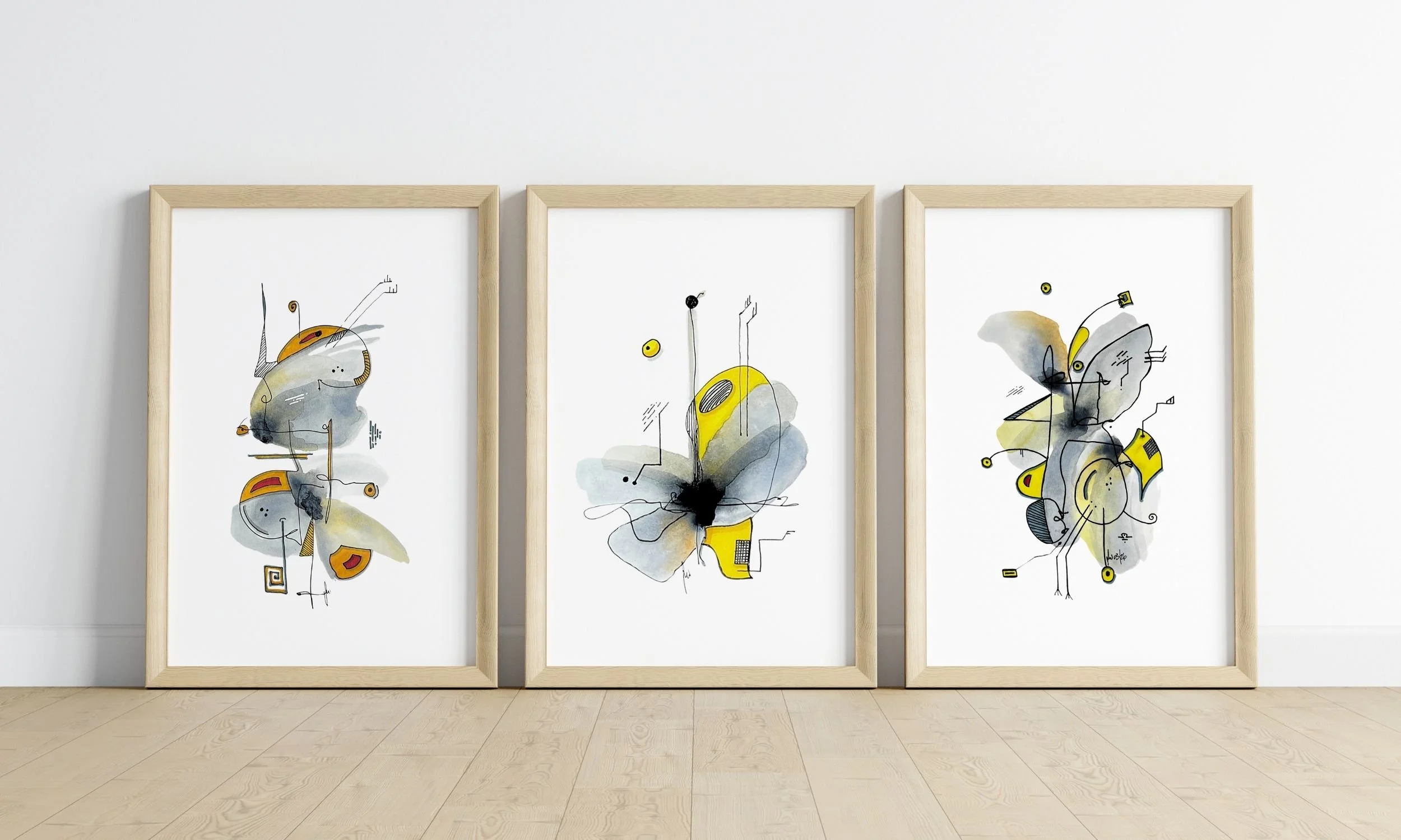 Three framed abstract watercolor and ink paintings from the Hybrid Collection in grey, yellow, and black tones, displayed in natural wood frames on a light wooden floor — original mixed media art by Olaf Wollmerstedt