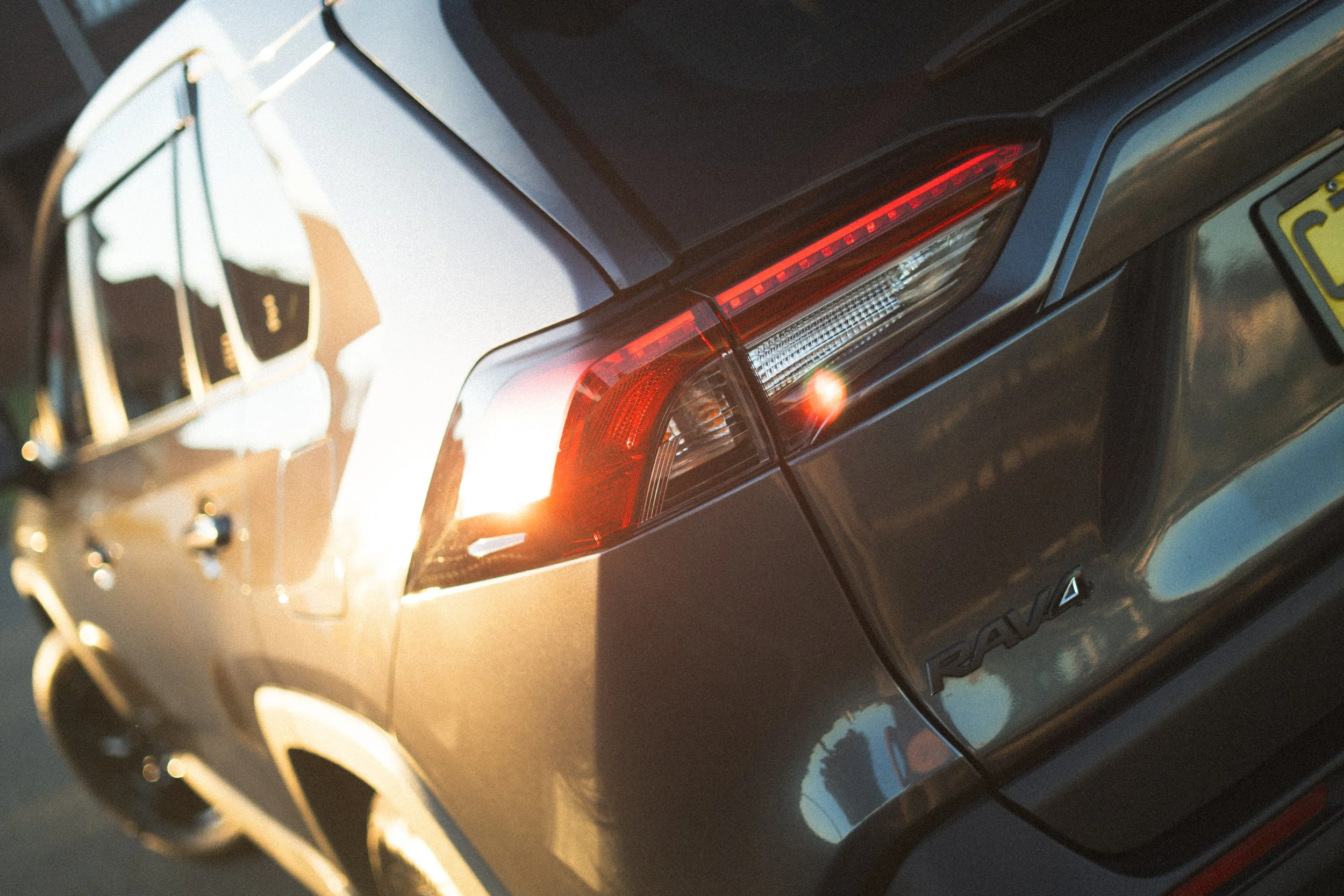 Close-up of the rear taillight and part of the trunk of a black luxury car, with blurred reflections and sunlight.
