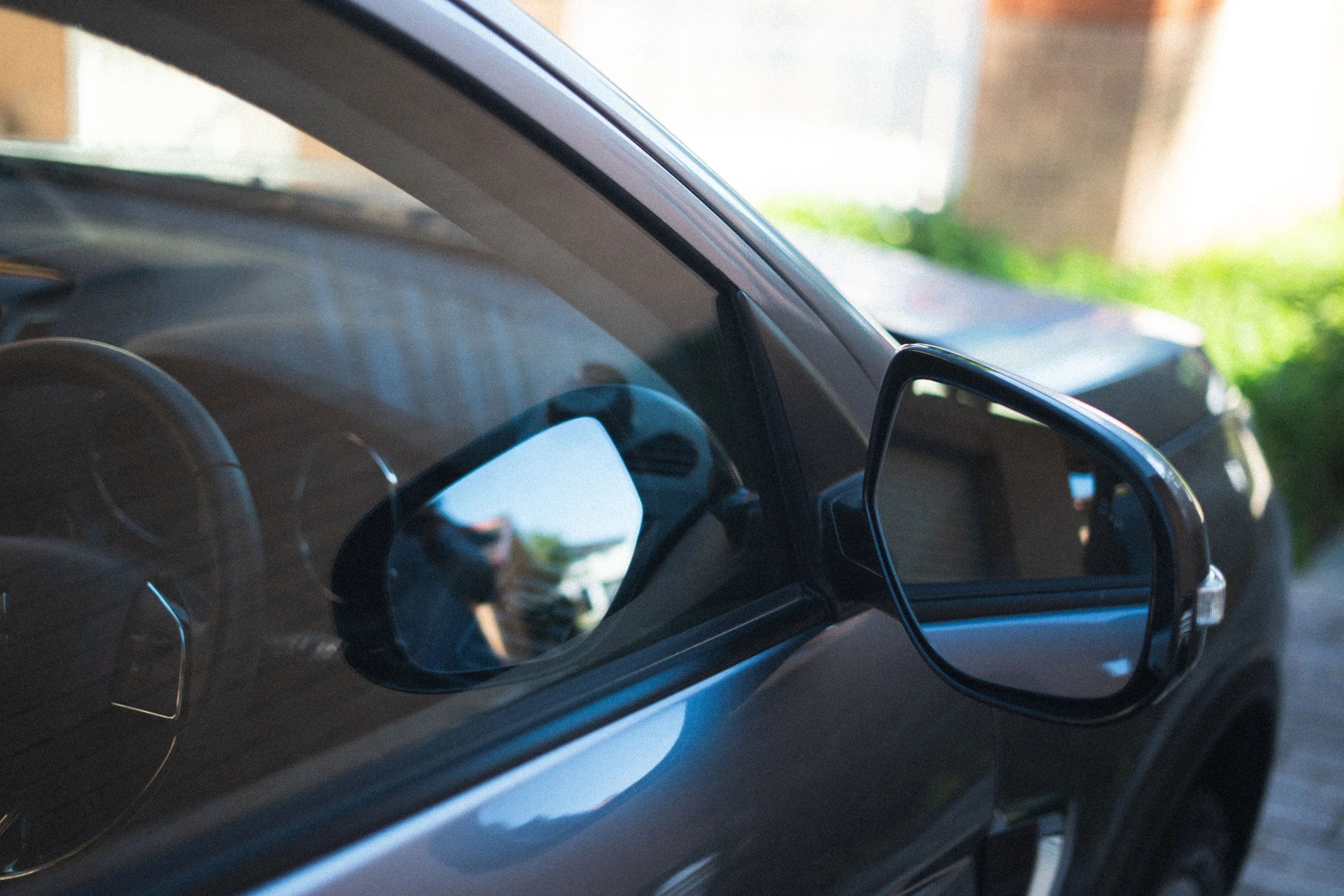 Close-up of a modern car's interior, showing the steering wheel and part of the dashboard and side mirror, with a background of green grass and blurred outdoor scenery.